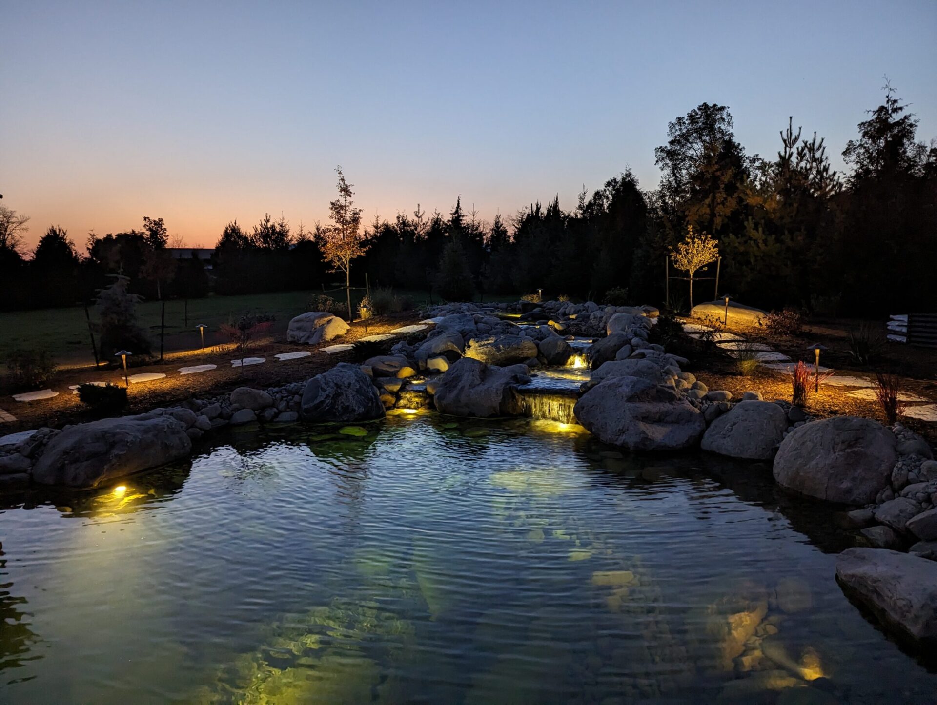 A tranquil garden scene at twilight features illuminated rocks and a peaceful pond surrounded by trees, creating a serene, picturesque landscape.