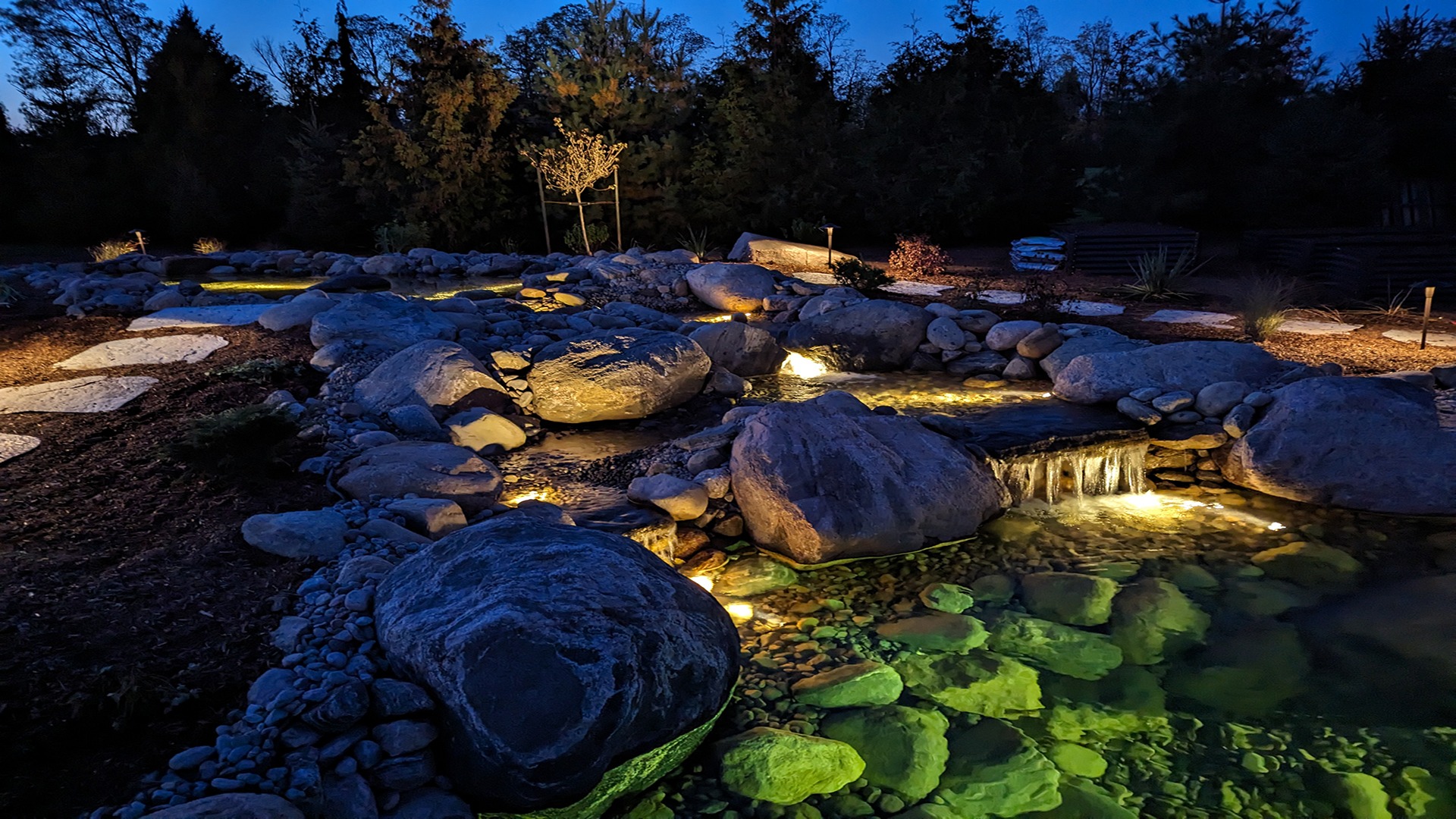 Illuminated rock garden at dusk; features small waterfall, glowing water, and stepping stones, surrounded by trees and ambient evening lighting. Peaceful ambiance.