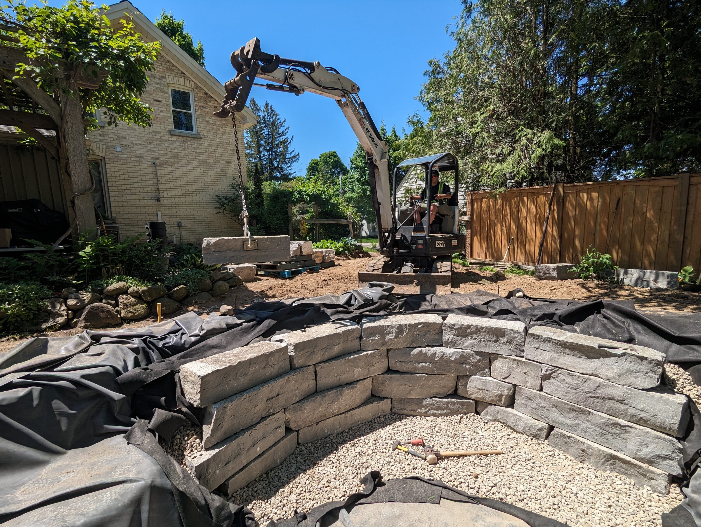 A person operates an excavator, placing large stones in a backyard. A brick house and wooden fence are visible under a clear sky.