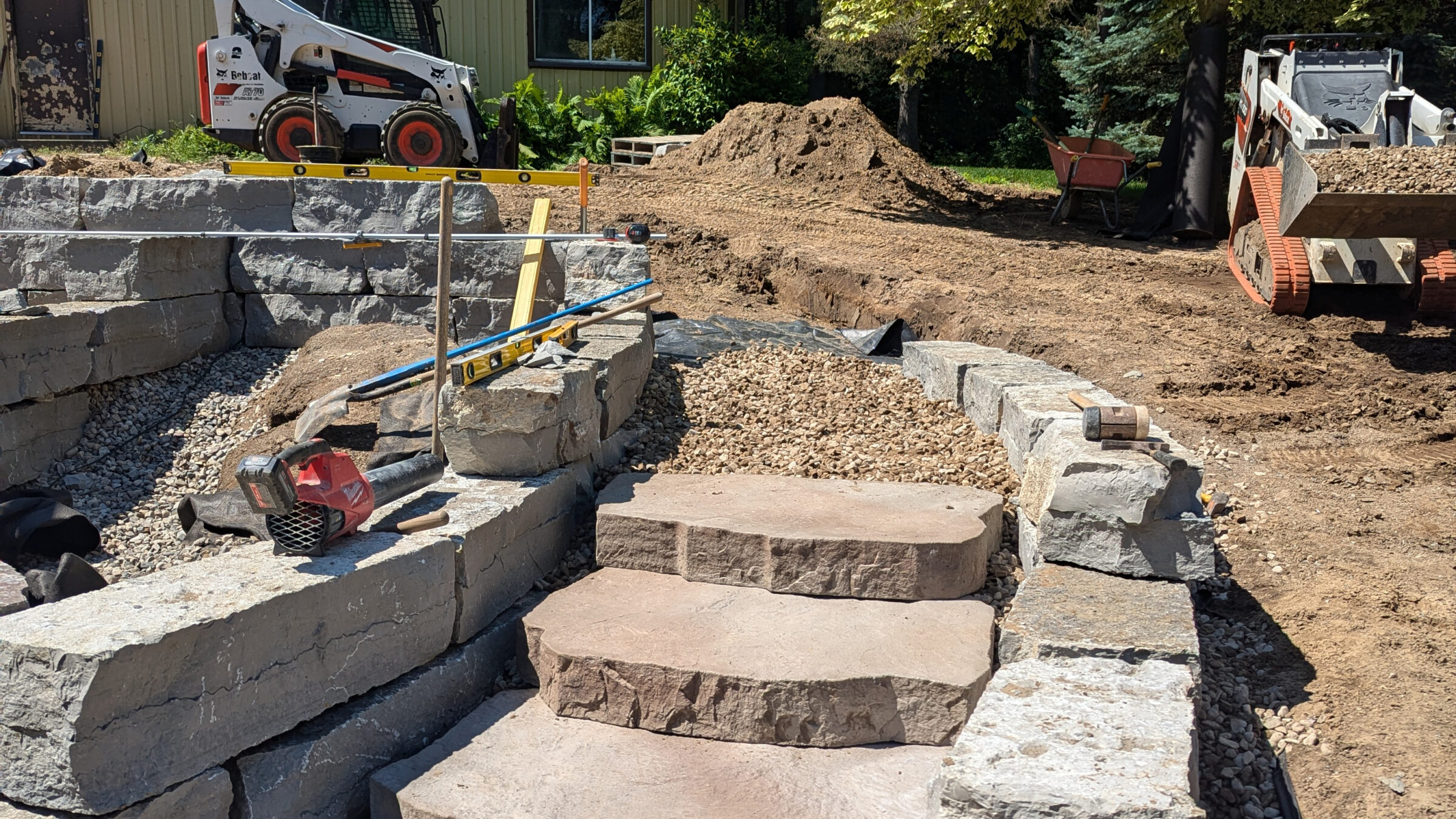 Construction site with machinery, stone steps, and retaining walls. Dirt piles and tools are scattered around, with a building visible in the background.