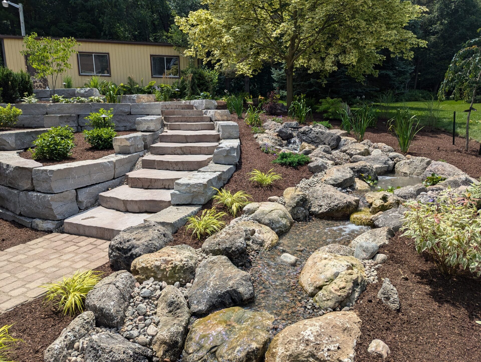 A landscaped garden features stone steps, a meandering stream, rocks, and lush greenery, adjacent to a yellow building and shaded by trees.