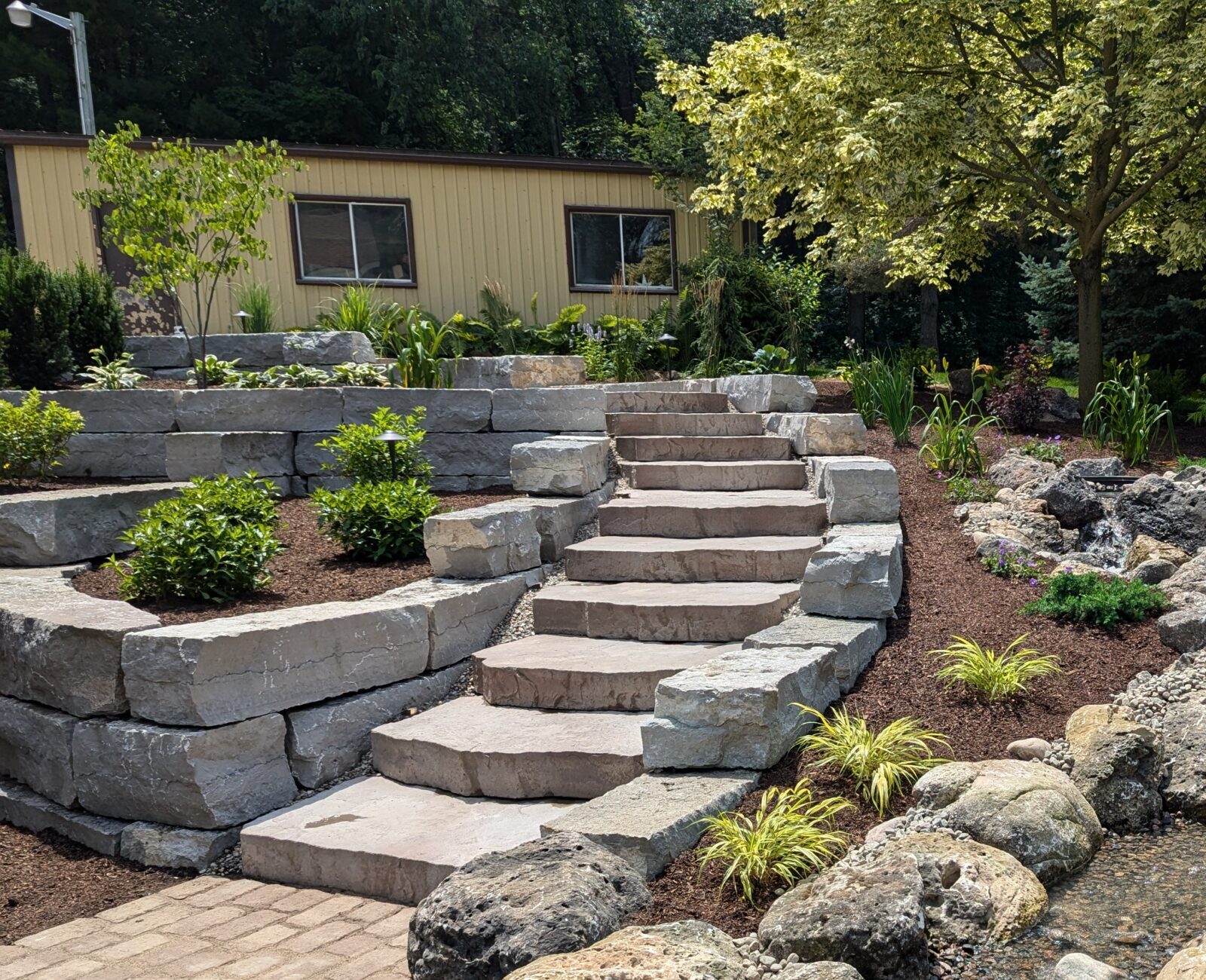 A landscaped garden features stone steps, a meandering stream, rocks, and lush greenery, adjacent to a yellow building and shaded by trees.