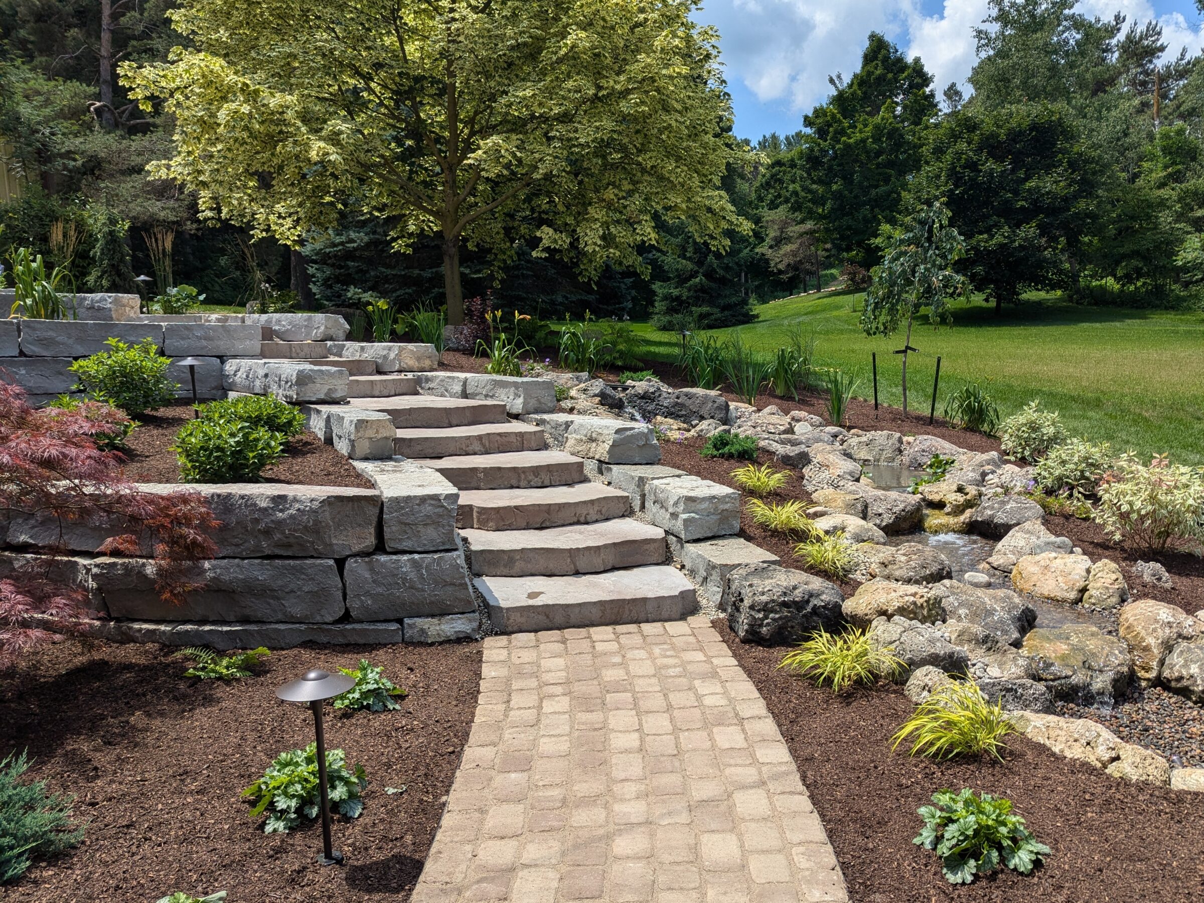 A garden path with stone steps, landscaped rocks, and plants leads up to a tree. Lush grass and trees in the background.