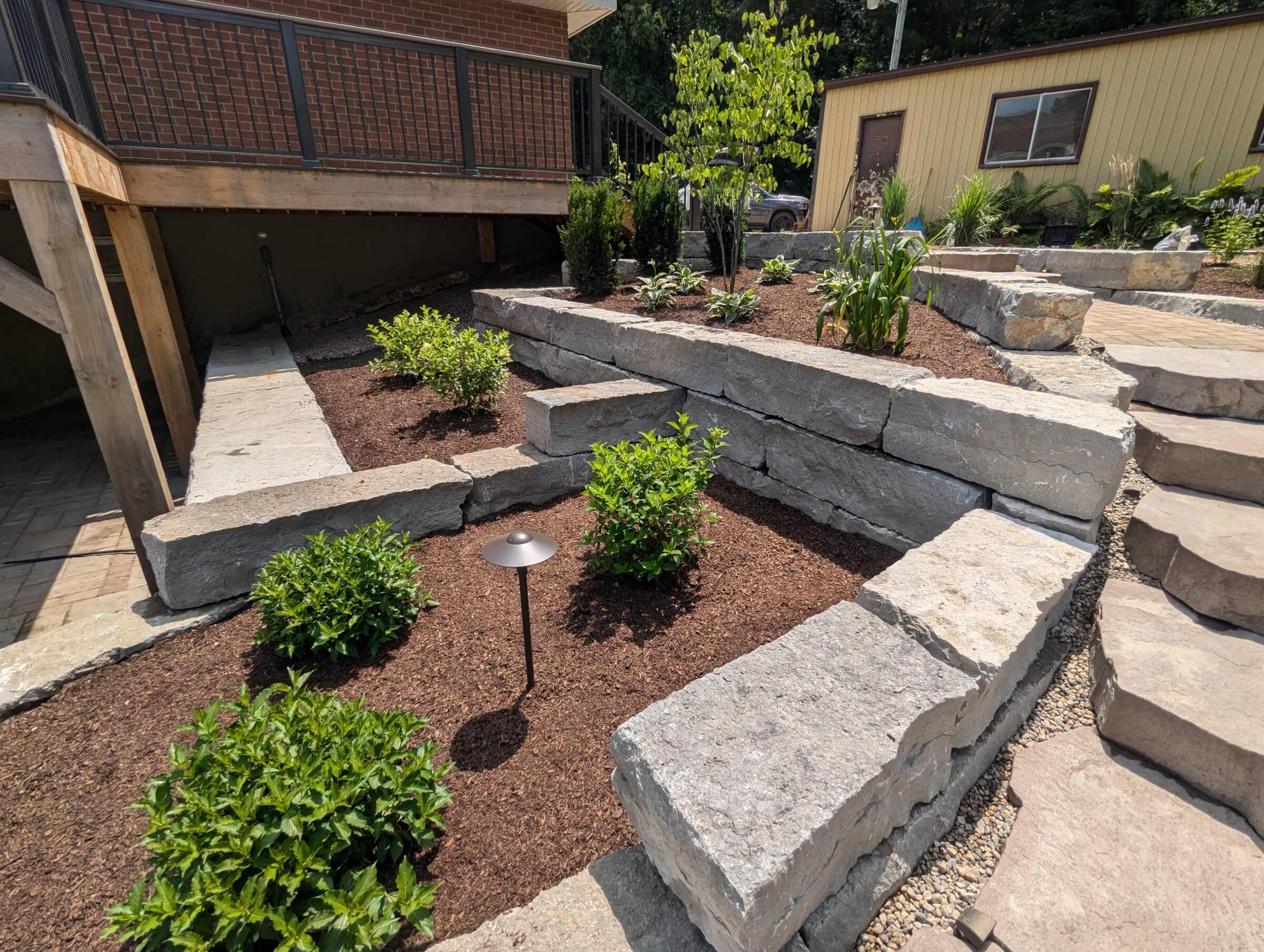 Terraced stone garden with lush green plants, mulch, and steps leading to a wooden deck with a railing. Small building nearby.