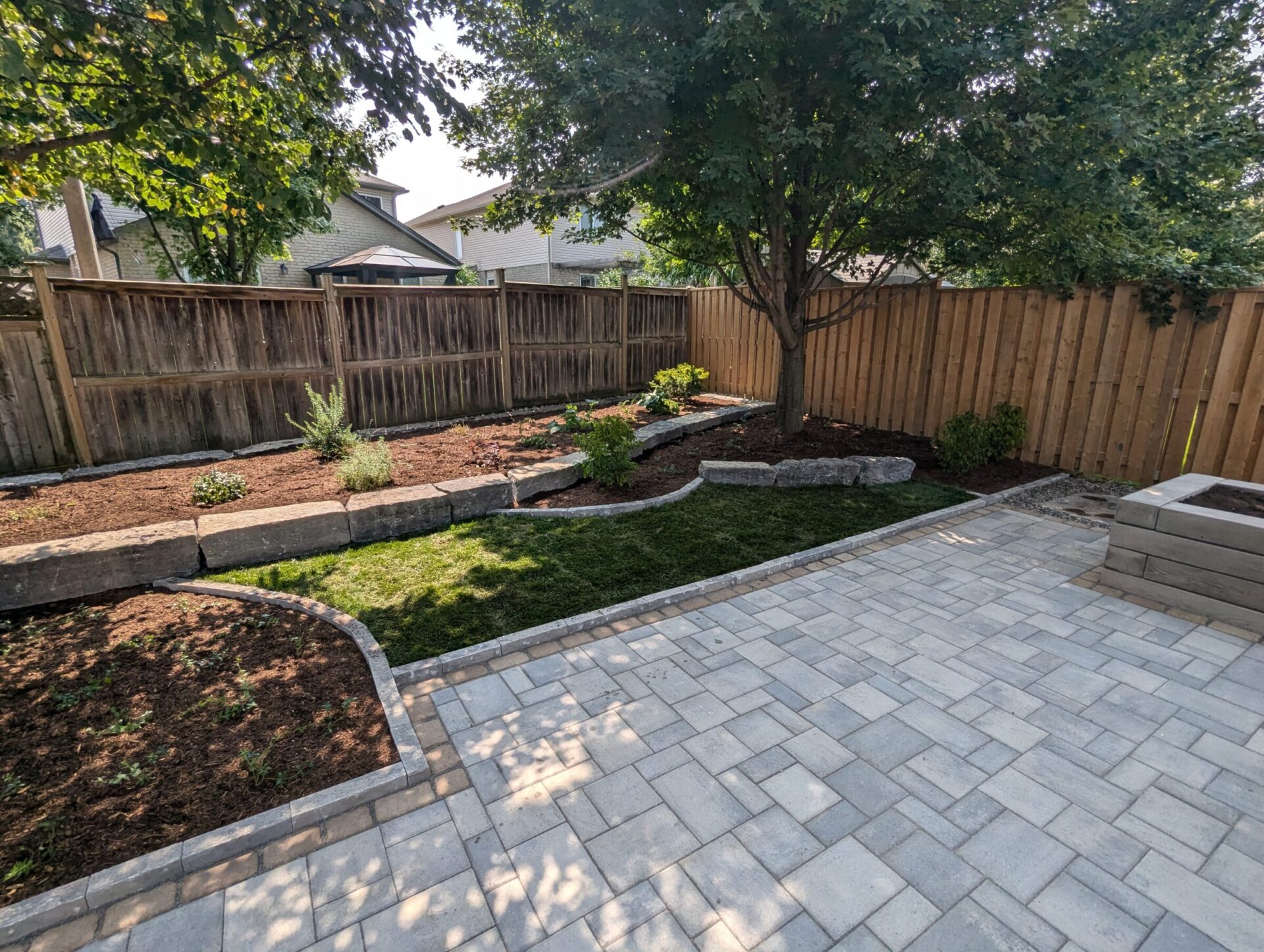 A landscaped backyard features a paved patio, garden beds, and a tree. Wooden fence surrounds the space with neighboring houses visible.