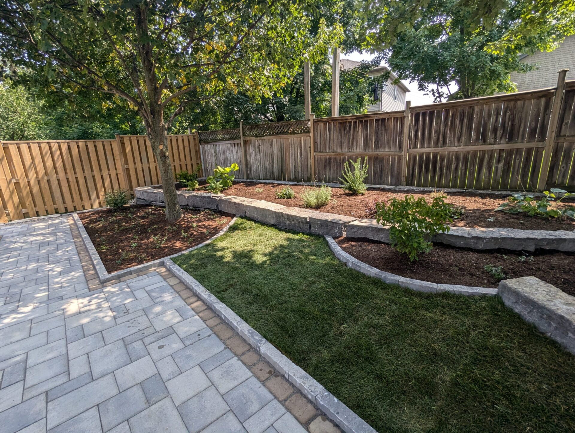 A neatly landscaped backyard with a stone patio, tree, and wooden fence, surrounded by mulch beds and green grass.