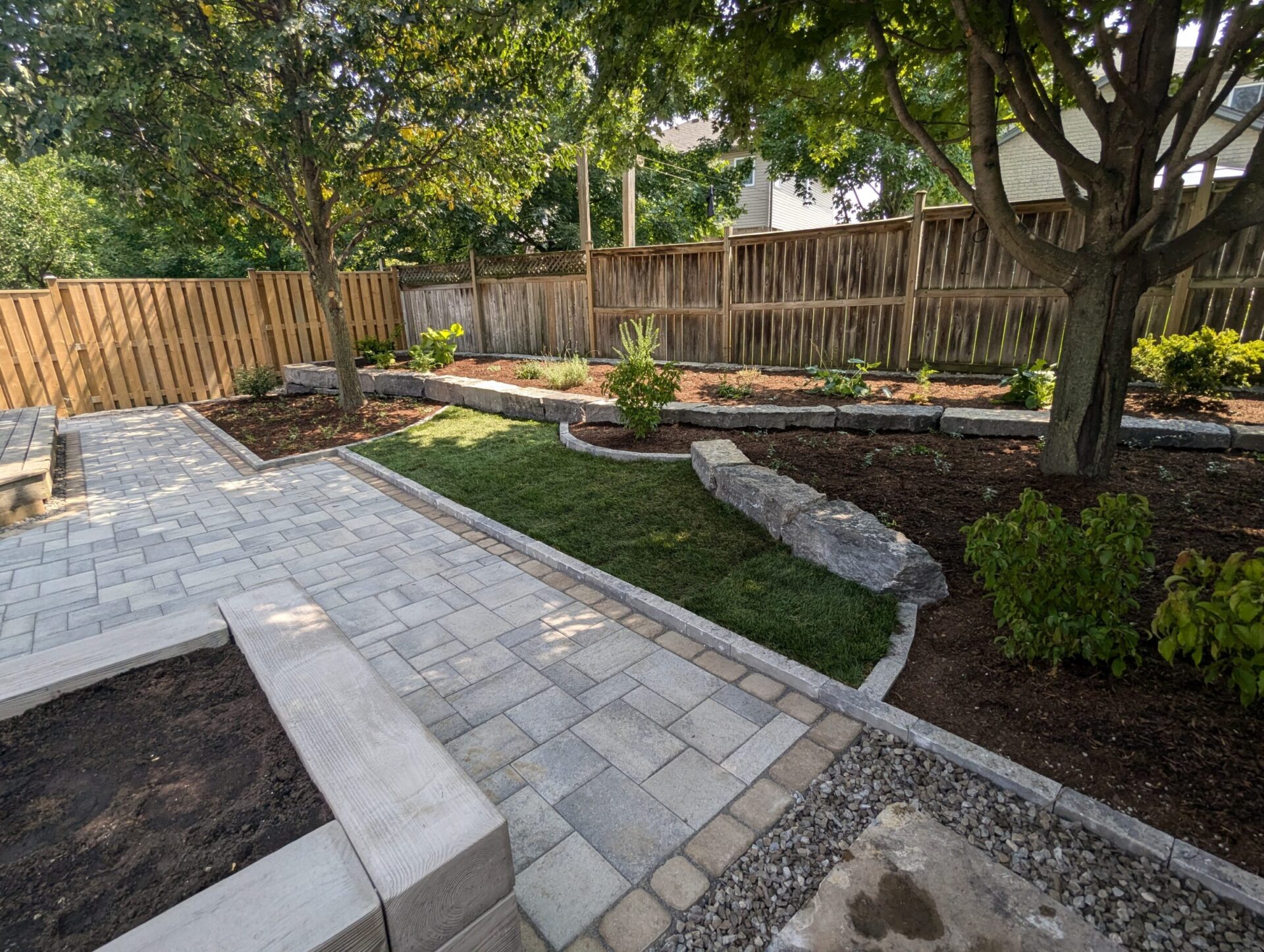 A landscaped backyard with stone pathways, wooden benches, trees, and a wooden fence, enhanced by neatly arranged plants and shrubs.