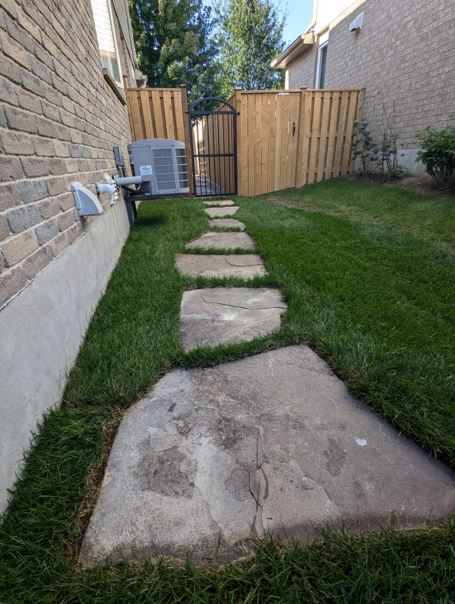 Stone path between two brick houses leads to a wooden gate, surrounded by grass and air conditioning unit visible.