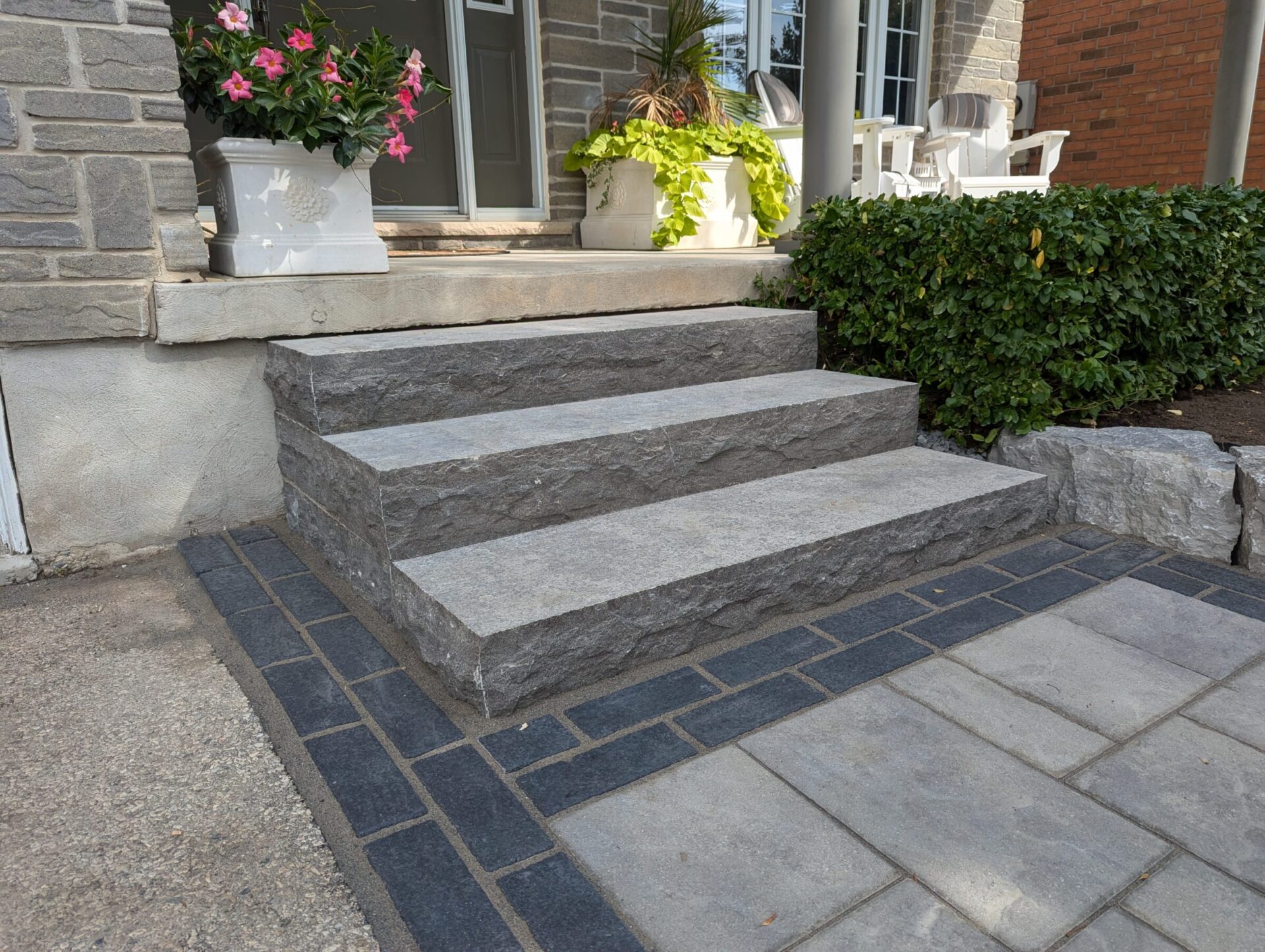 Stone steps lead to a front porch with potted flowers and greenery beside brick and concrete walls.