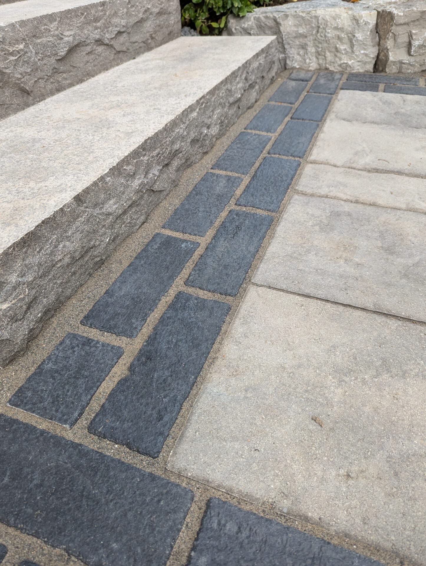 Close-up of stone steps with a bordering pathway. The design features dark and light bricks arranged in a pattern. Greenery visible in background.