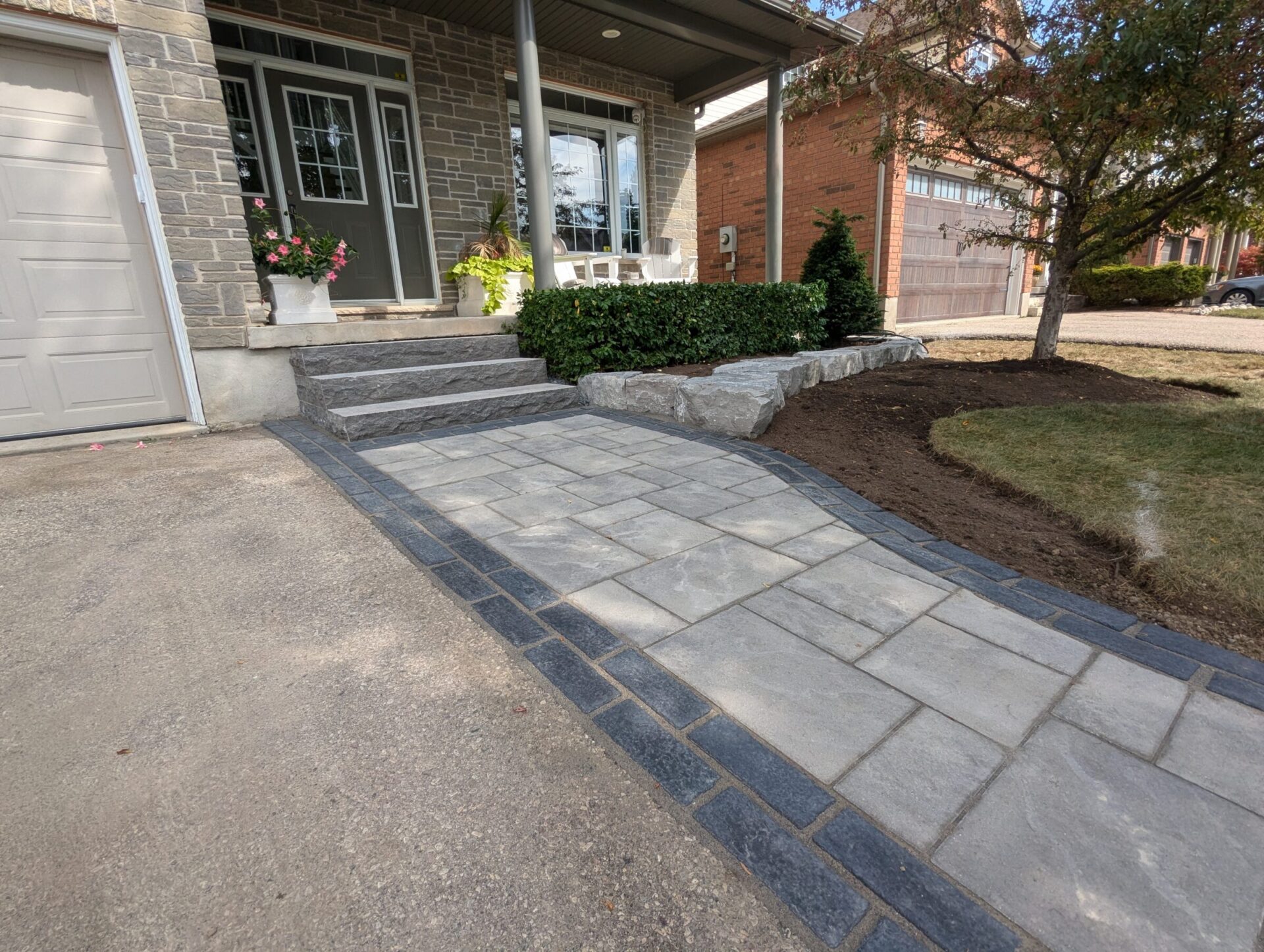 A residential entrance with a stone pathway, steps, potted plants, and landscaped garden near a garage, with brick and stone exterior walls.