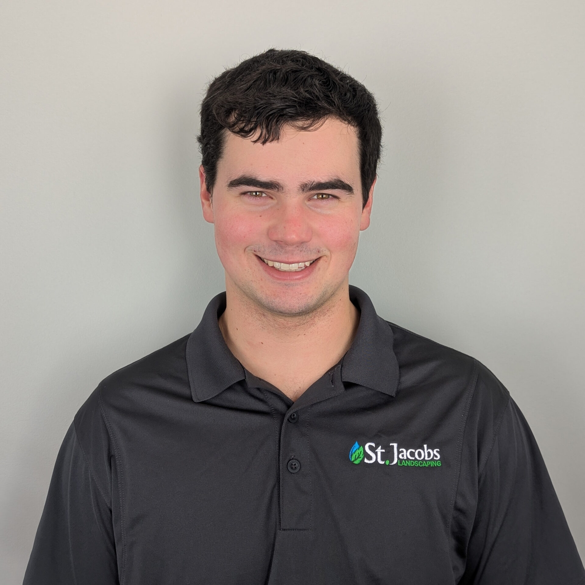 A person wearing a dark polo shirt with "St. Jacobs Landscaping" logo, standing against a plain, light-colored background, smiling warmly.