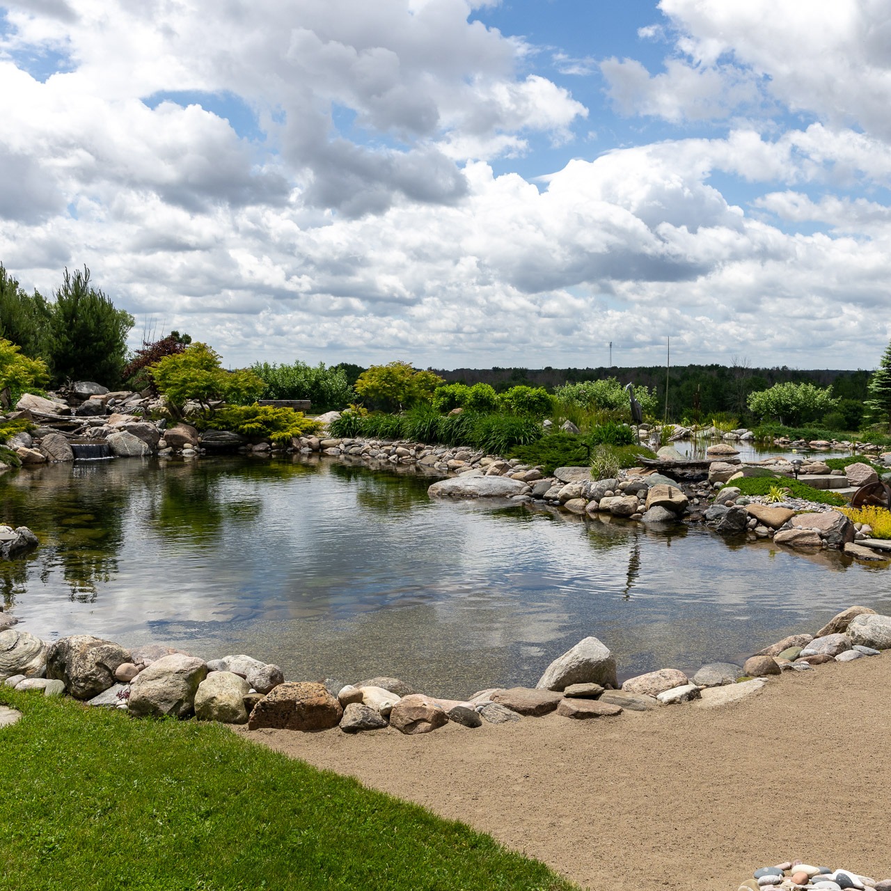 A serene garden with a tranquil pond, surrounded by rocks and lush greenery under a partly cloudy sky. No people or buildings visible.