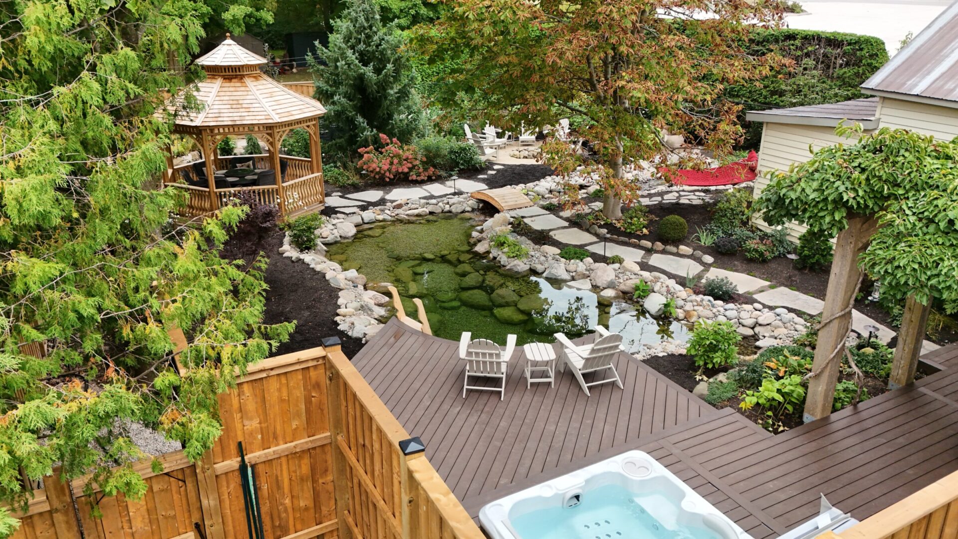 A serene backyard features a gazebo, koi pond, wooden deck with chairs, and a hot tub, surrounded by lush trees and plants.