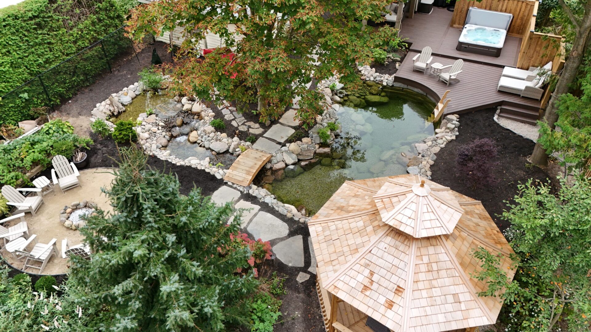 Aerial view of a landscaped garden featuring a pond, wooden gazebo, seating area, and jacuzzi on a wooden deck surrounded by greenery.