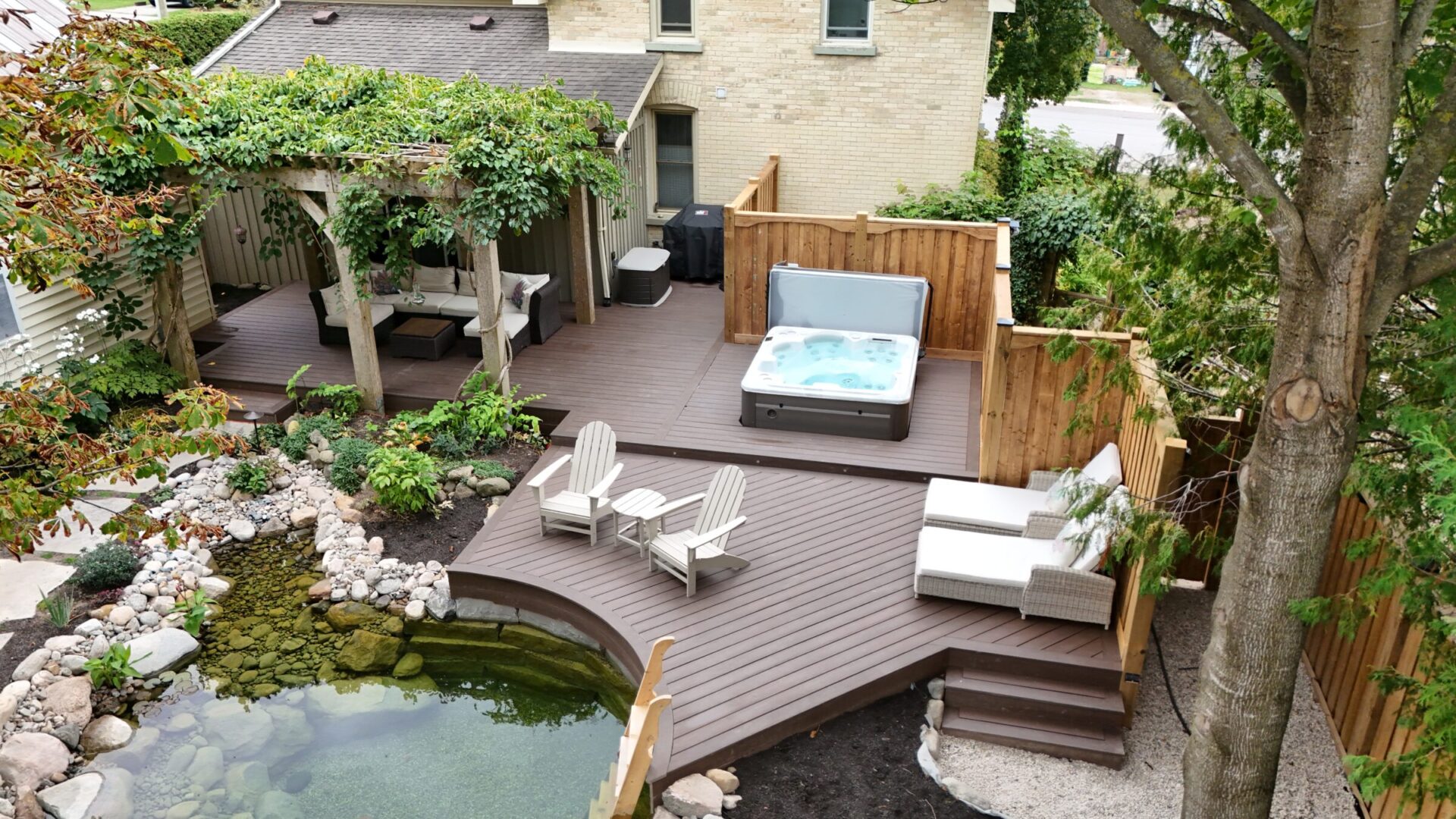 A serene backyard features a wooden deck, hot tub, pergola with seating area, and landscaped pond, surrounded by lush greenery and fencing.