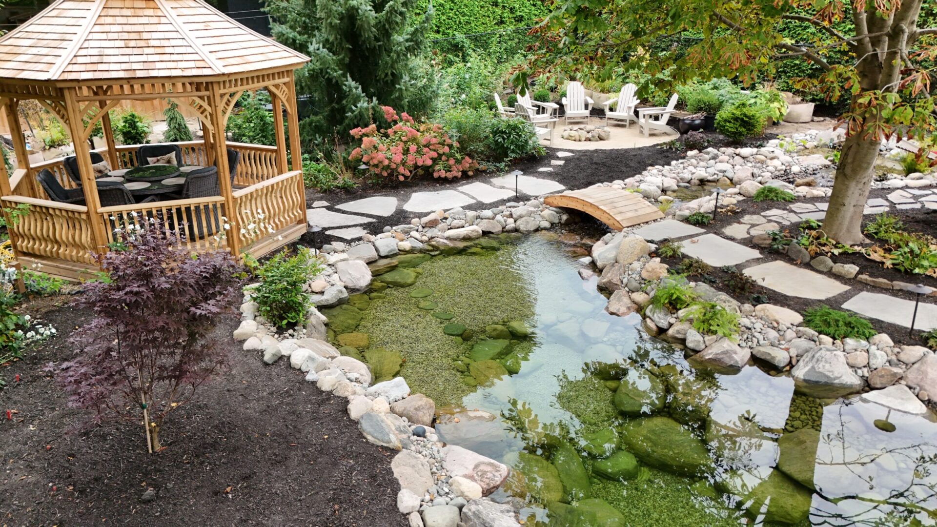 A serene garden features a wooden gazebo, small pond with rocks, a tree, and adirondack chairs, creating a peaceful outdoor setting.