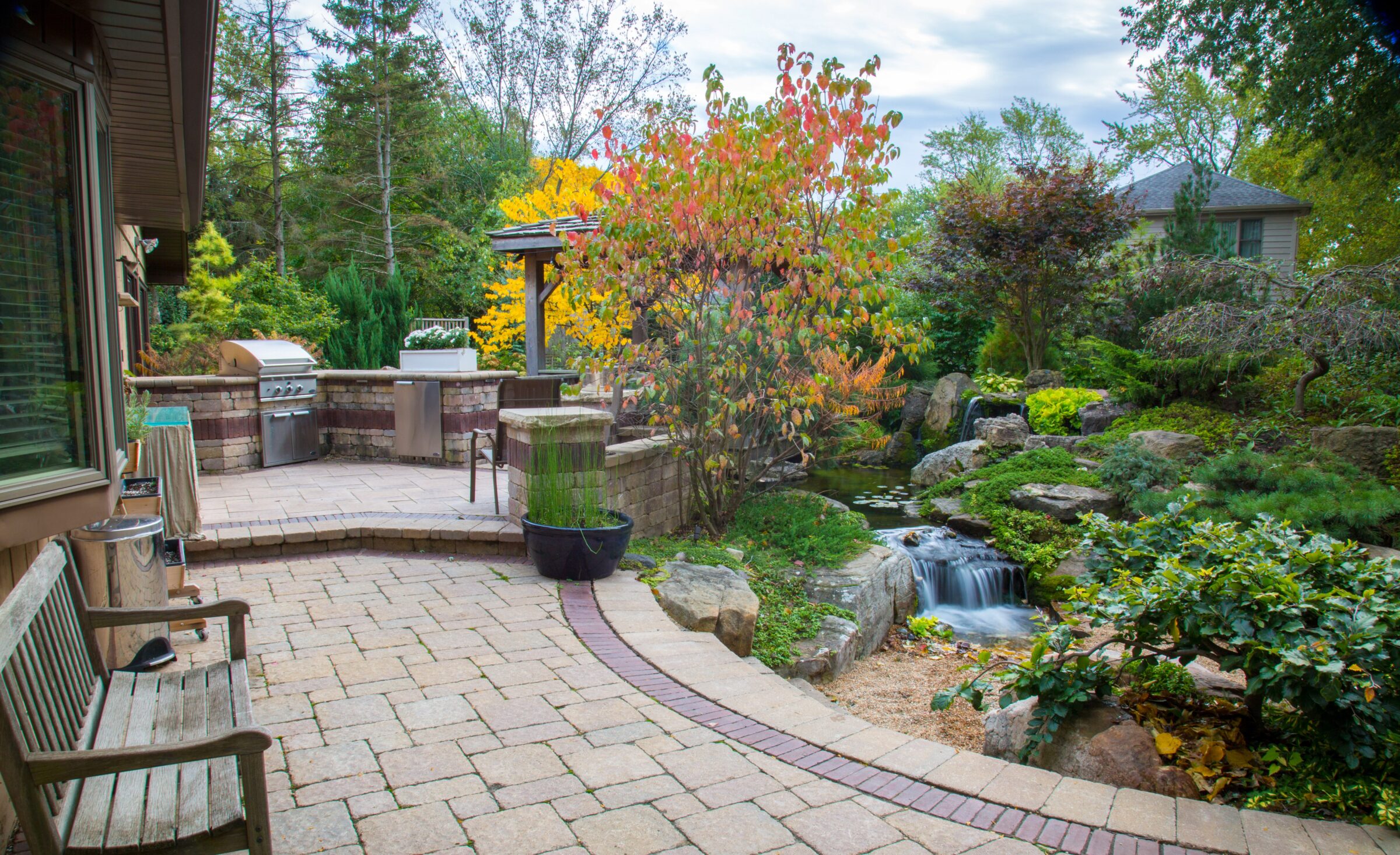 Stone patio with outdoor grill, surrounded by lush garden and small waterfall. Wooden bench and colorful trees create a serene atmosphere.
