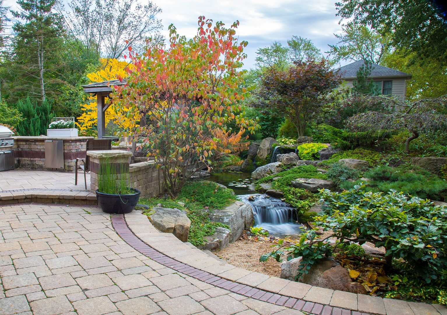 A vibrant garden with a patio, barbecue, colorful trees, and a serene waterfall cascading into a small pond. Peaceful and inviting.