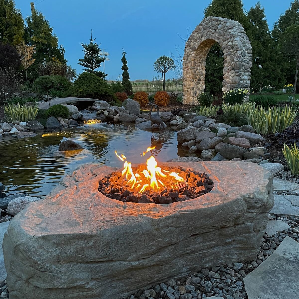 A stone fire pit glows beside a serene pond with a stone arch and lush greenery in the background, creating a tranquil setting.