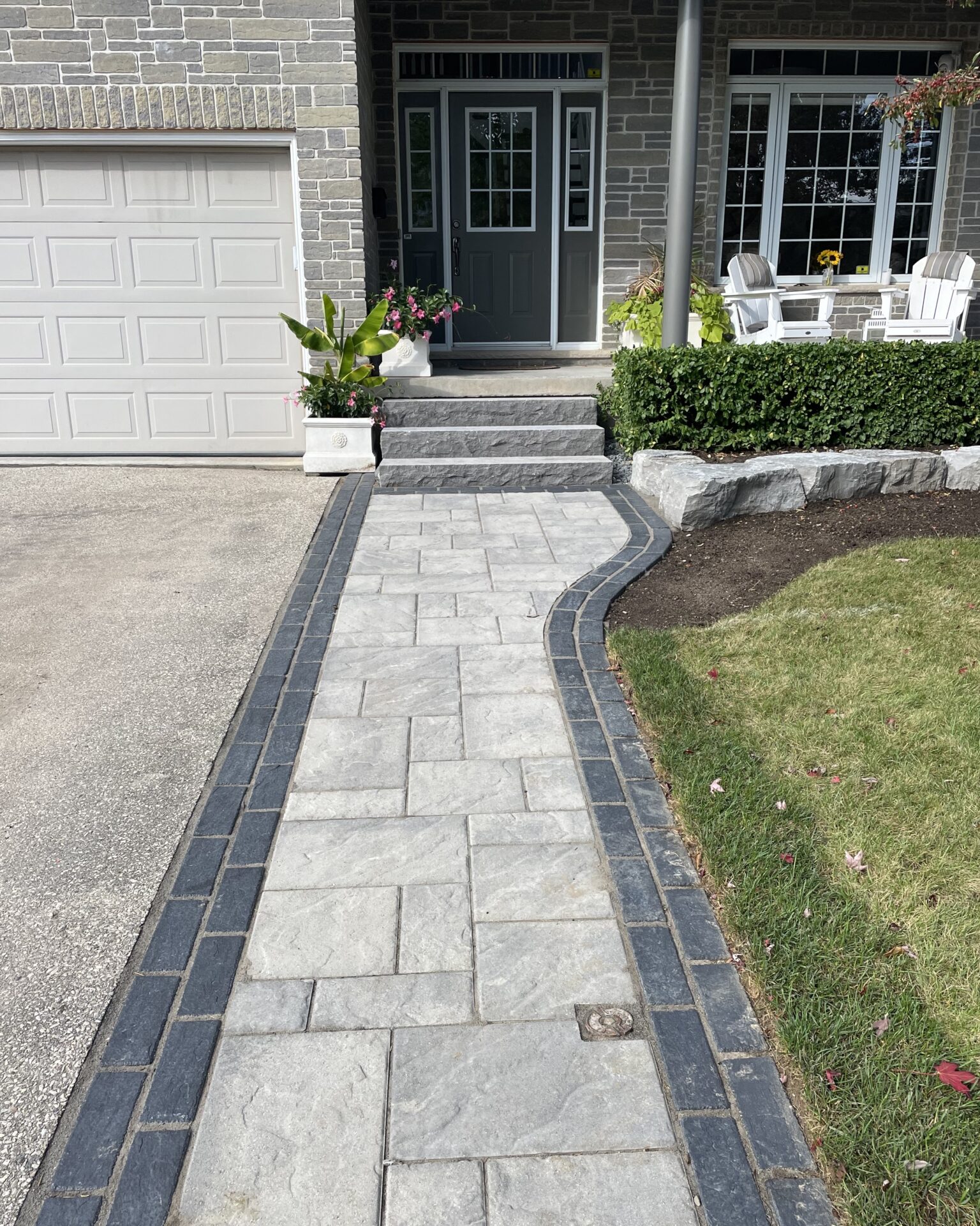 A neat stone pathway leads to a house with potted plants, bushes, and white chairs on a porch near a gray door.