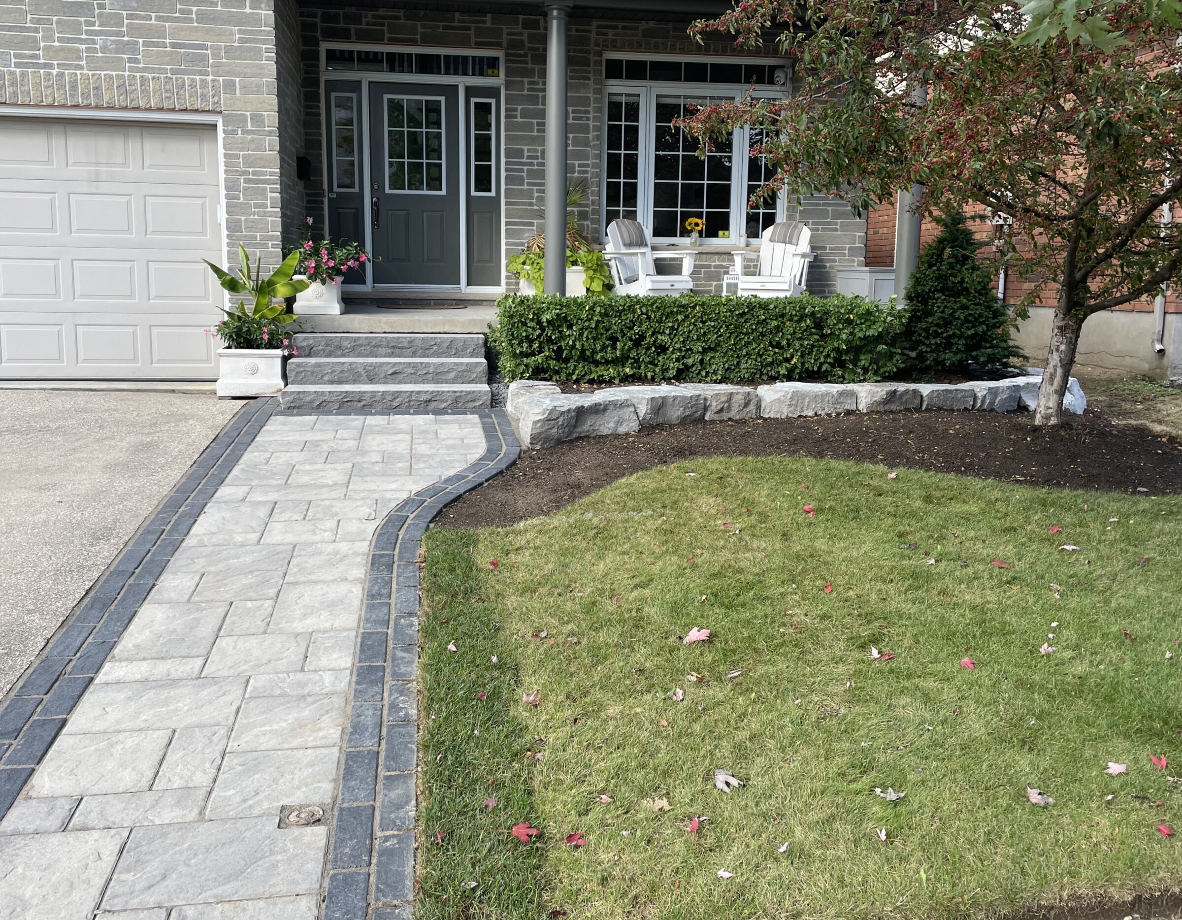 A paved walkway leads to a cozy porch with chairs and plants, surrounded by a manicured lawn and a small tree.