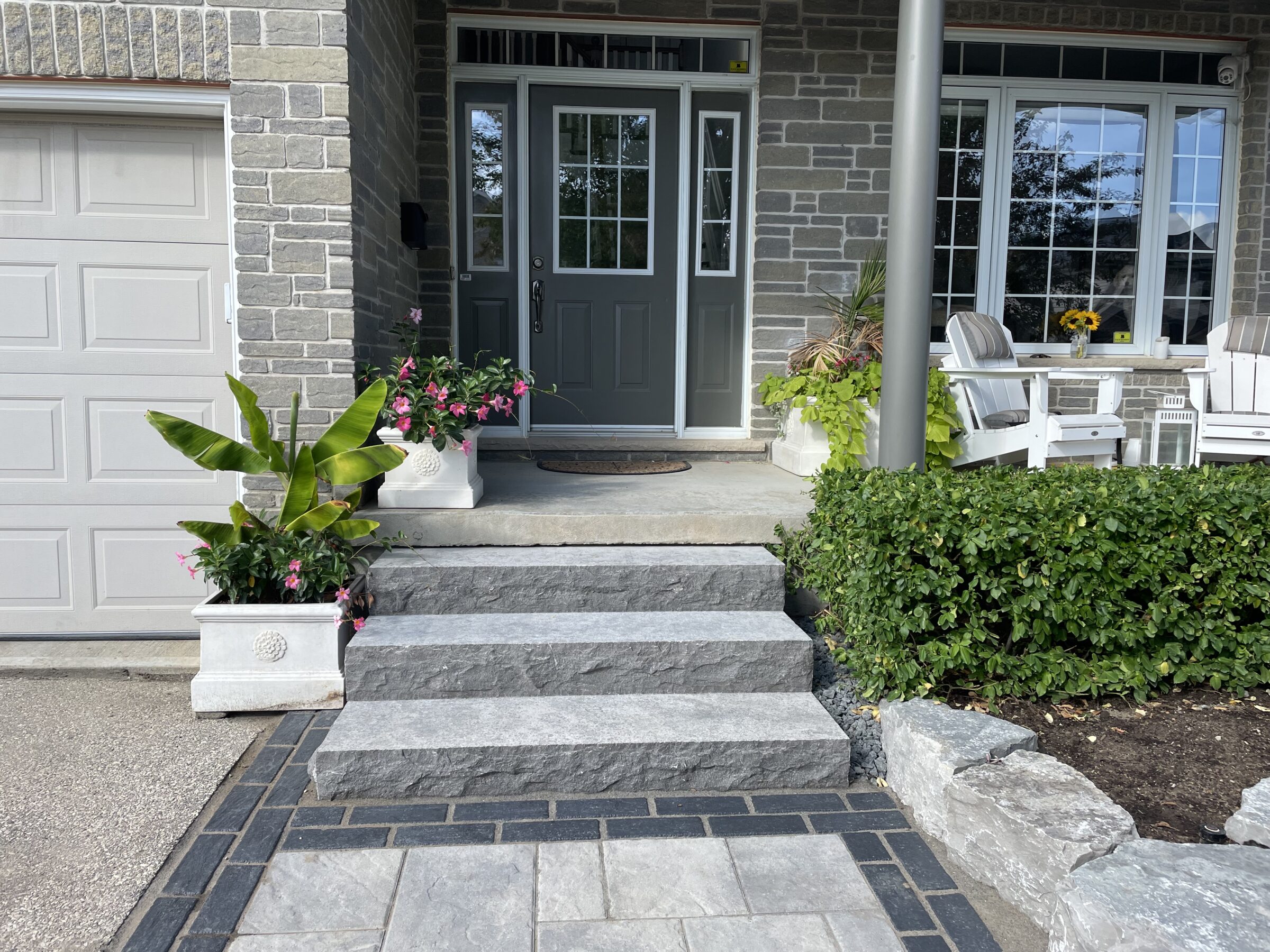 A modern house entrance with stone steps, planters, and Adirondack chairs on a porch. Nearby are green bushes and a gray garage door.