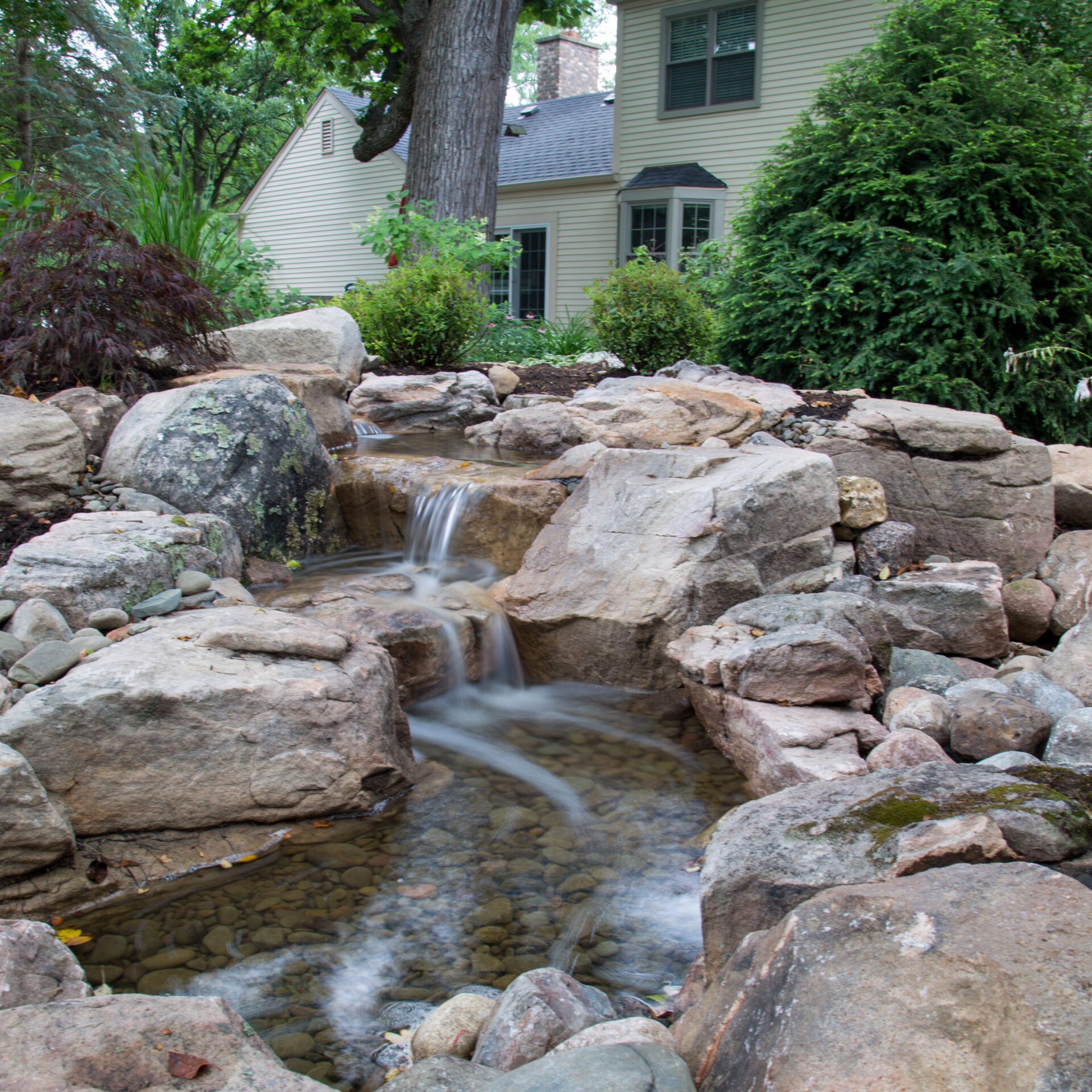 A serene garden waterfall flows over rocks, surrounded by lush greenery, with a charming house in the background, creating a peaceful ambiance.