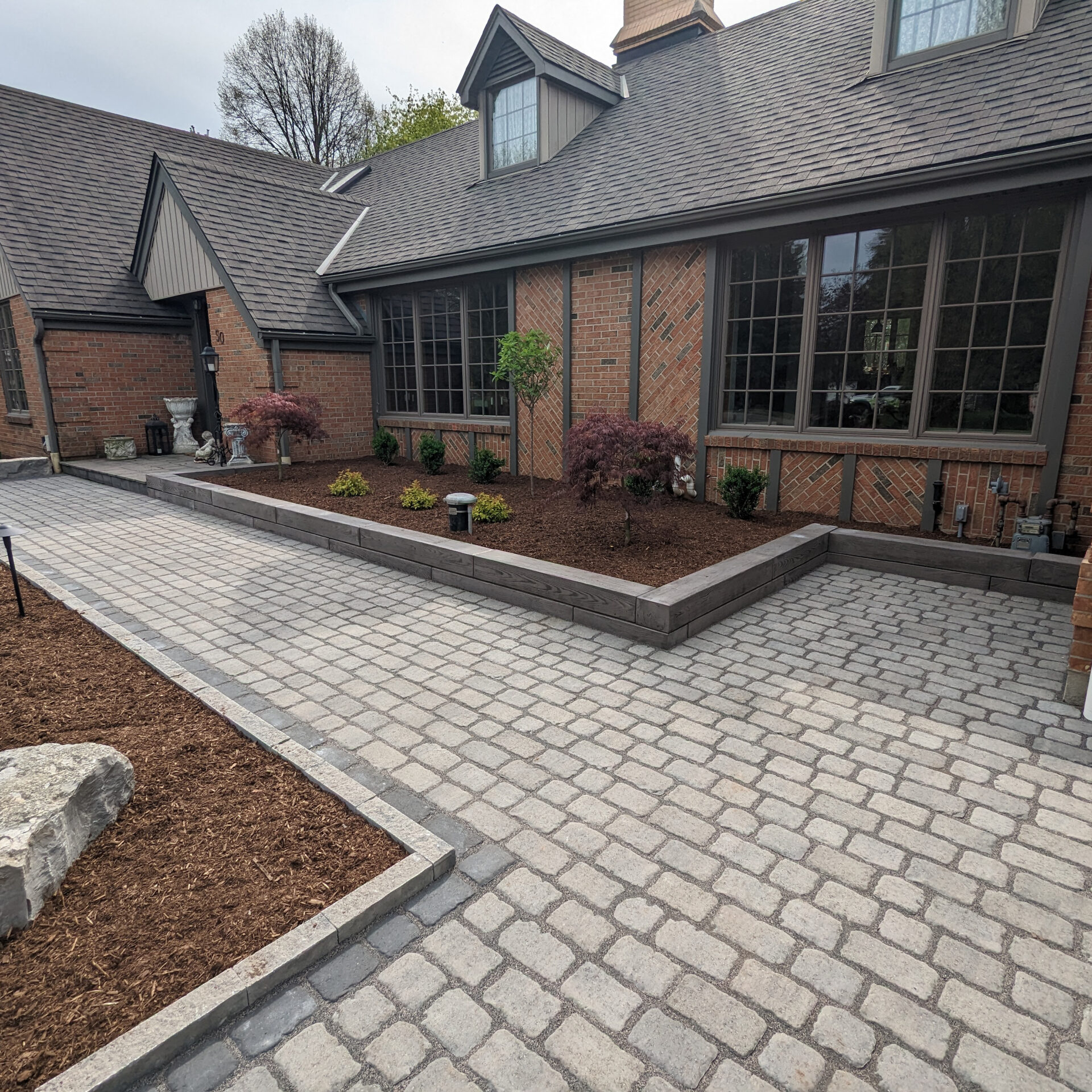 A brick house with large windows, a stone pathway, and a tidy garden area featuring small shrubs and neatly trimmed bushes.