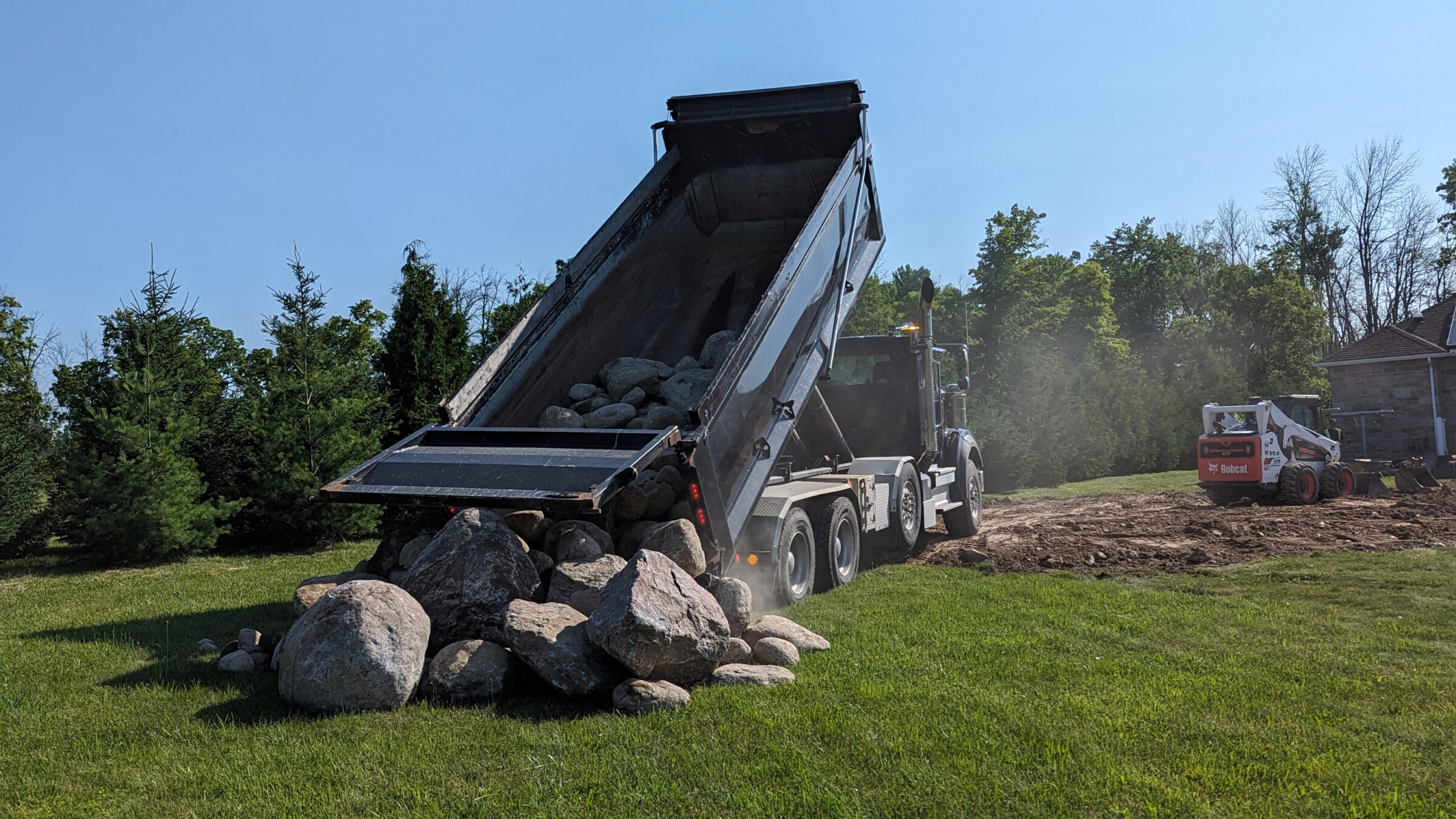 A dump truck unloads large rocks onto grass beside trees; a small loader operates nearby under a clear blue sky.