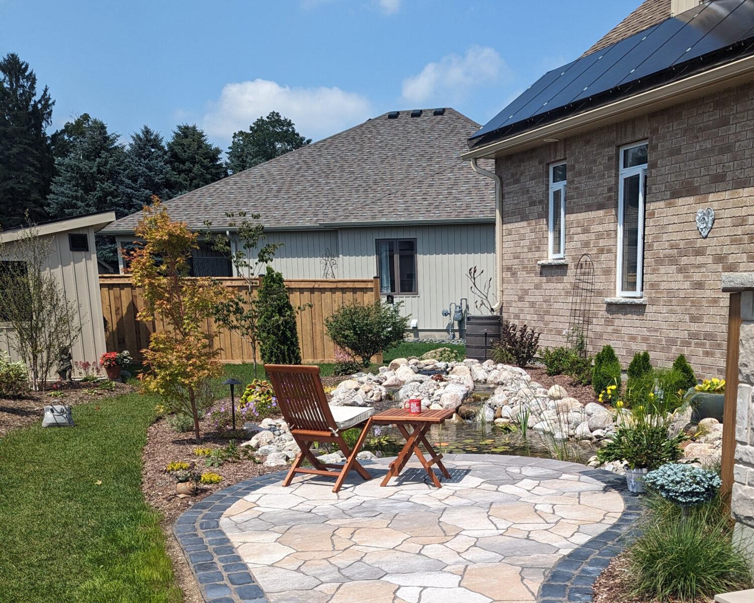 A paved patio with chairs and table, surrounded by a garden and steps, sits next to a brick house with a solar panel.