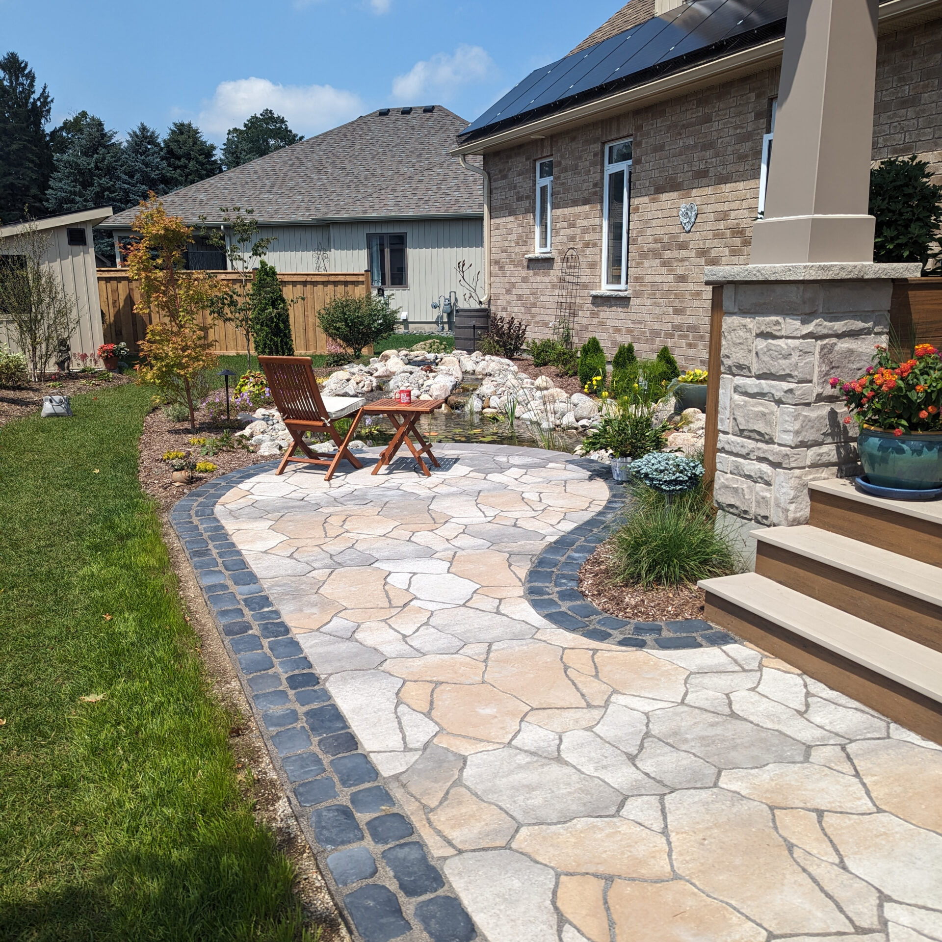 A paved patio with chairs and table, surrounded by a garden and steps, sits next to a brick house with a solar panel.