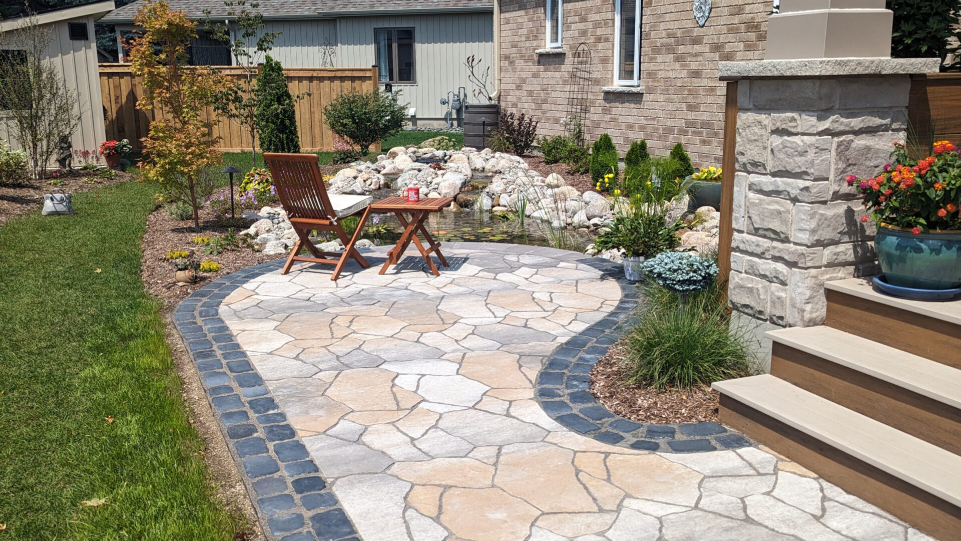 A tranquil backyard patio with stone pavers, surrounded by plants, features wooden chairs, a table, and a small pond with rocks.