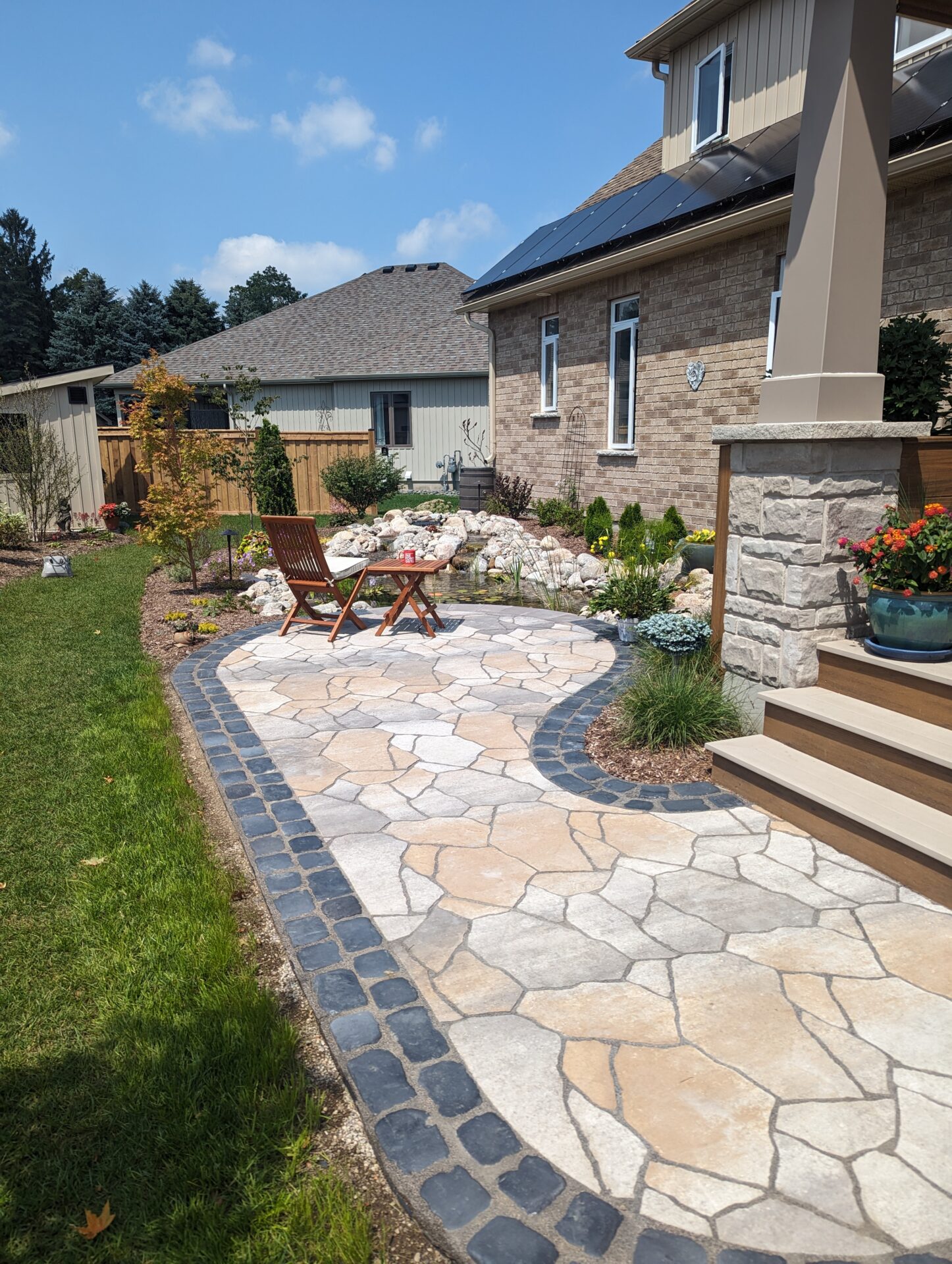 A peaceful backyard with a stone path and garden, featuring a pond and solar panels on a modern house. Two chairs are arranged outside.