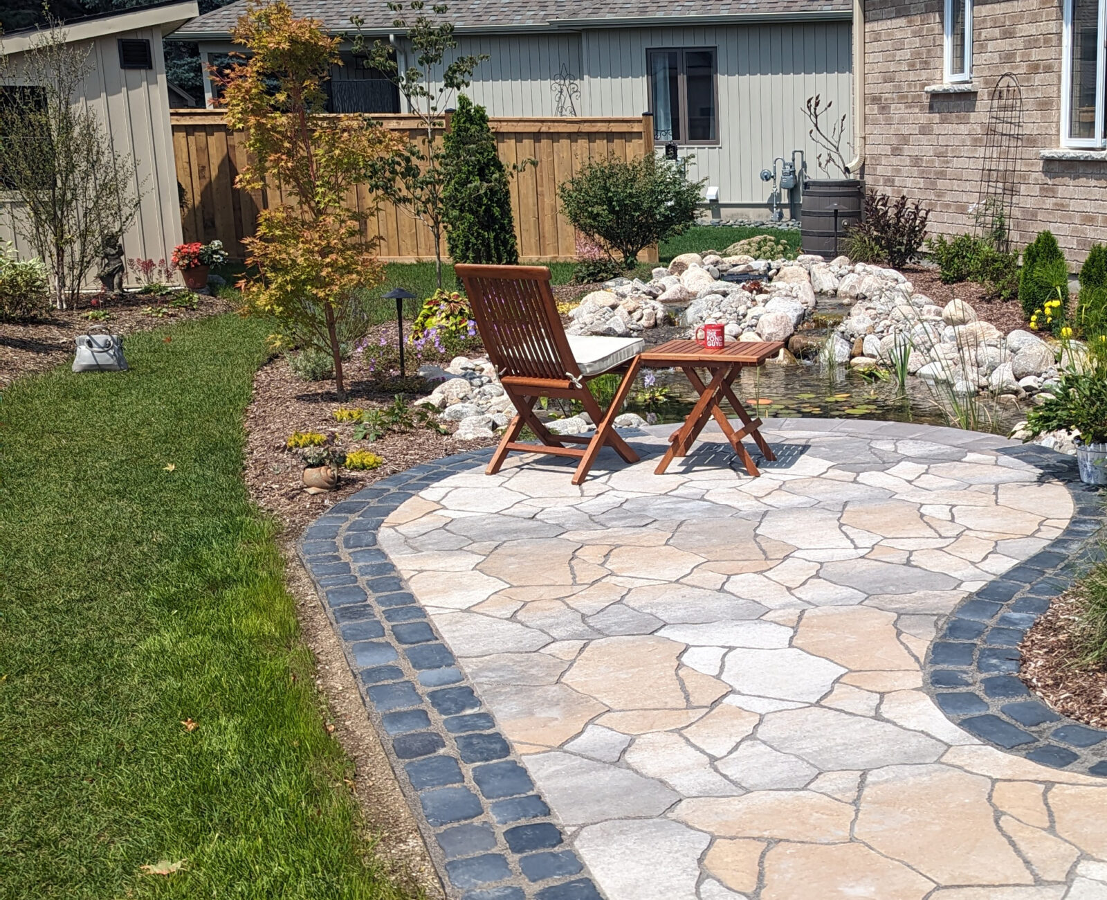 A tranquil backyard patio with stone pavers, surrounded by plants, features wooden chairs, a table, and a small pond with rocks.