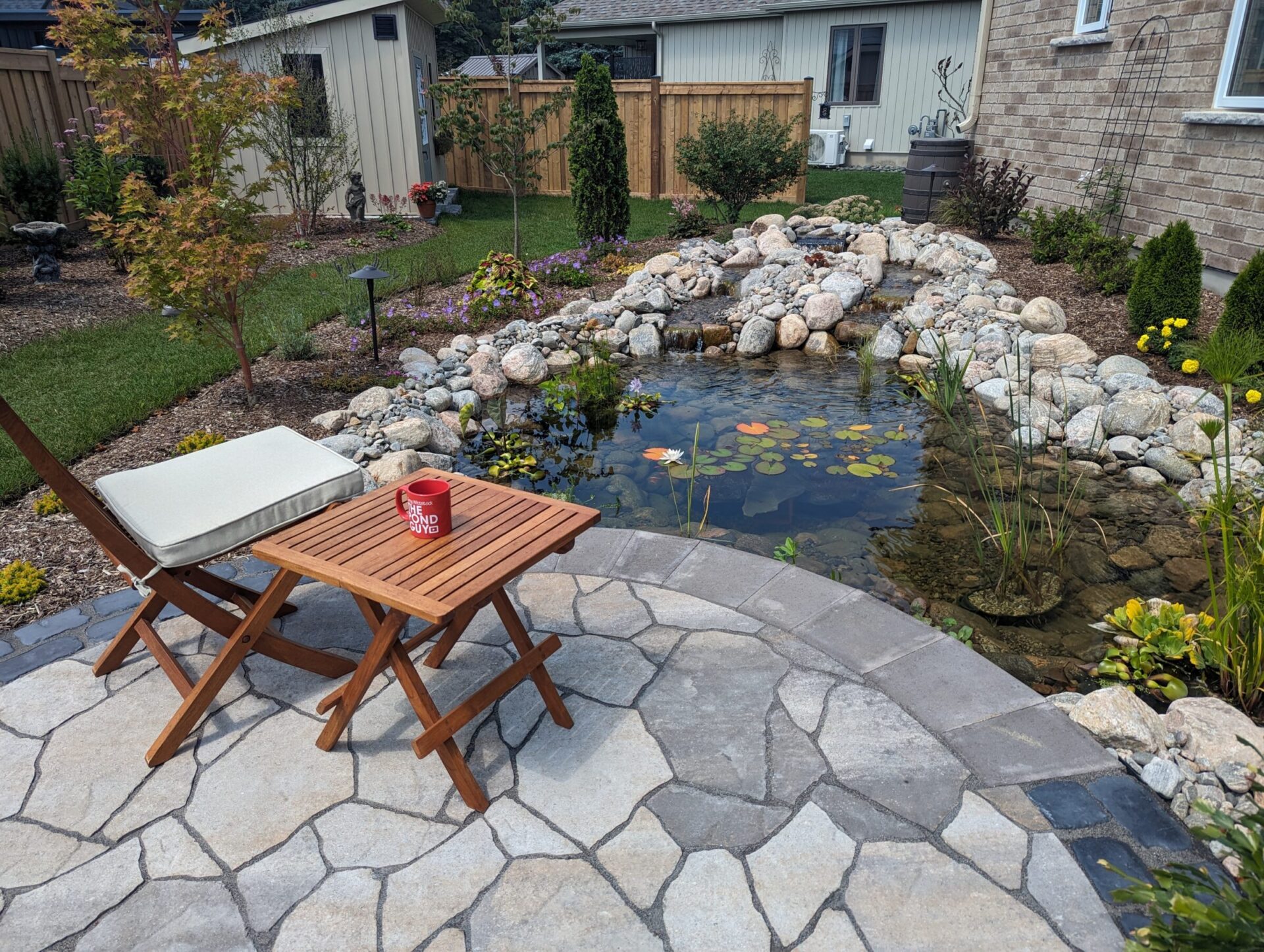 A garden with a stone patio, koi pond, chair, and table with a red mug. Surrounded by plants and a wooden fence.