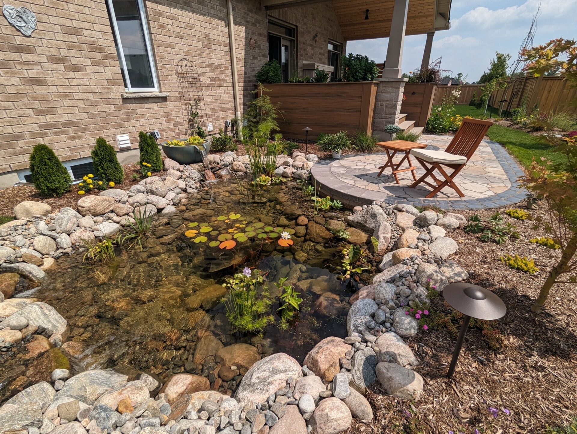 A peaceful garden features a small pond with lilies, surrounded by rocks, chairs on a patio, and a landscaped yard.