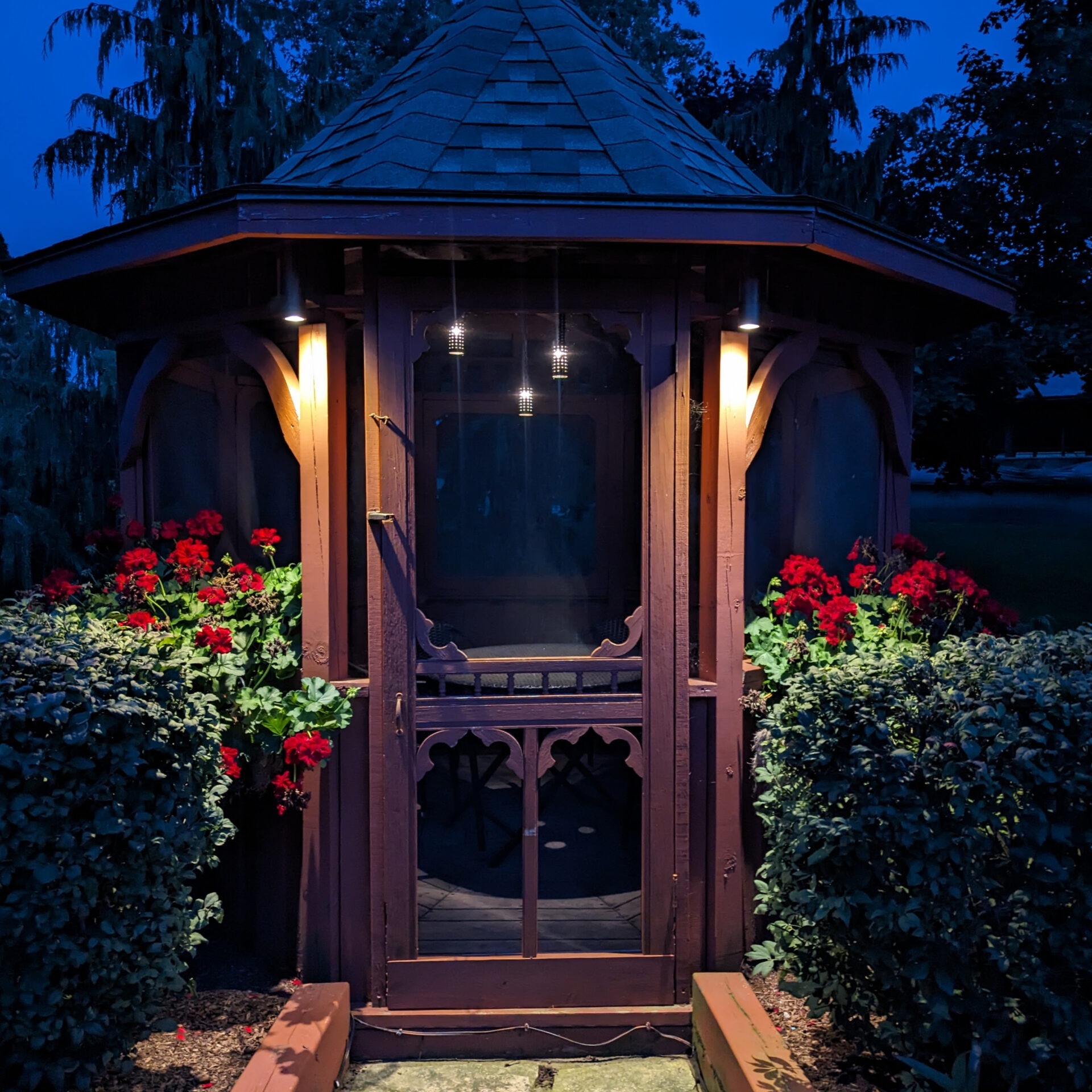 A small, illuminated wooden gazebo surrounded by red flowers and bushes, set against a backdrop of trees at dusk.