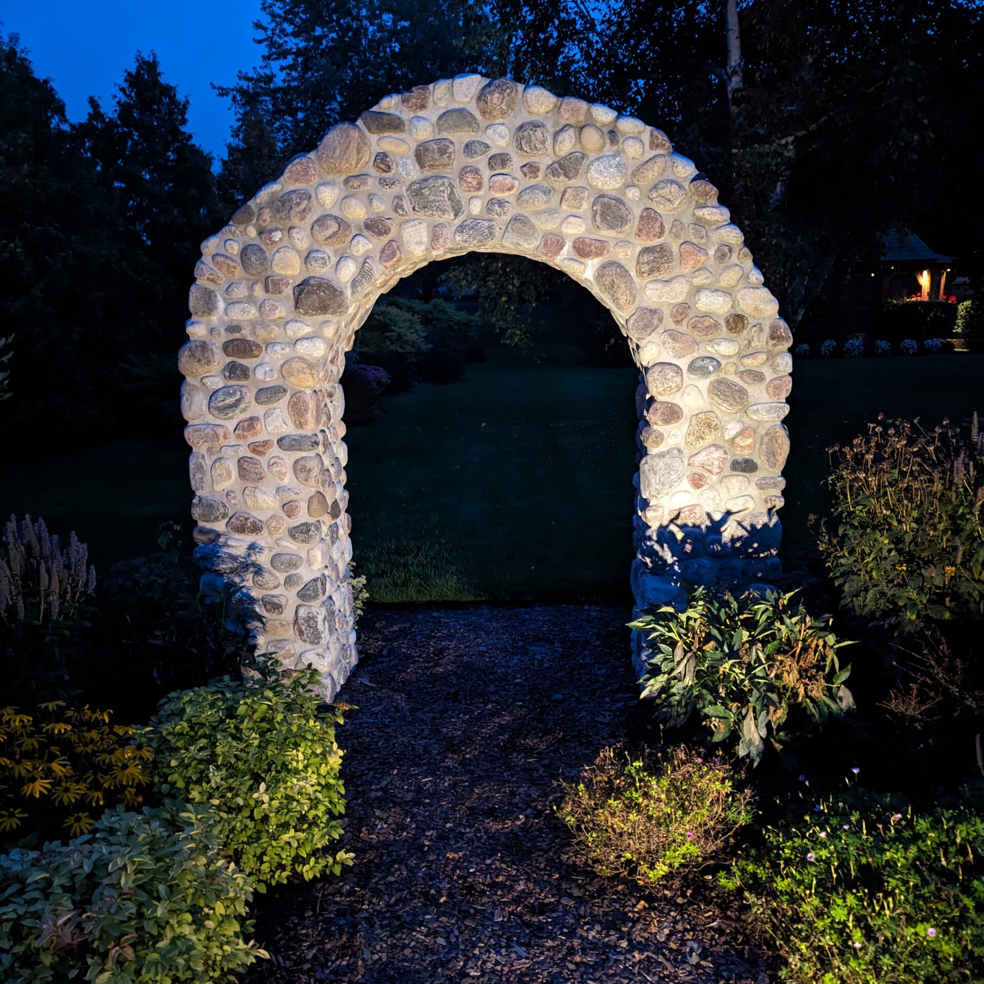 A stone archway in a garden is illuminated at dusk, surrounded by bushes and flowers, creating a tranquil evening scene.