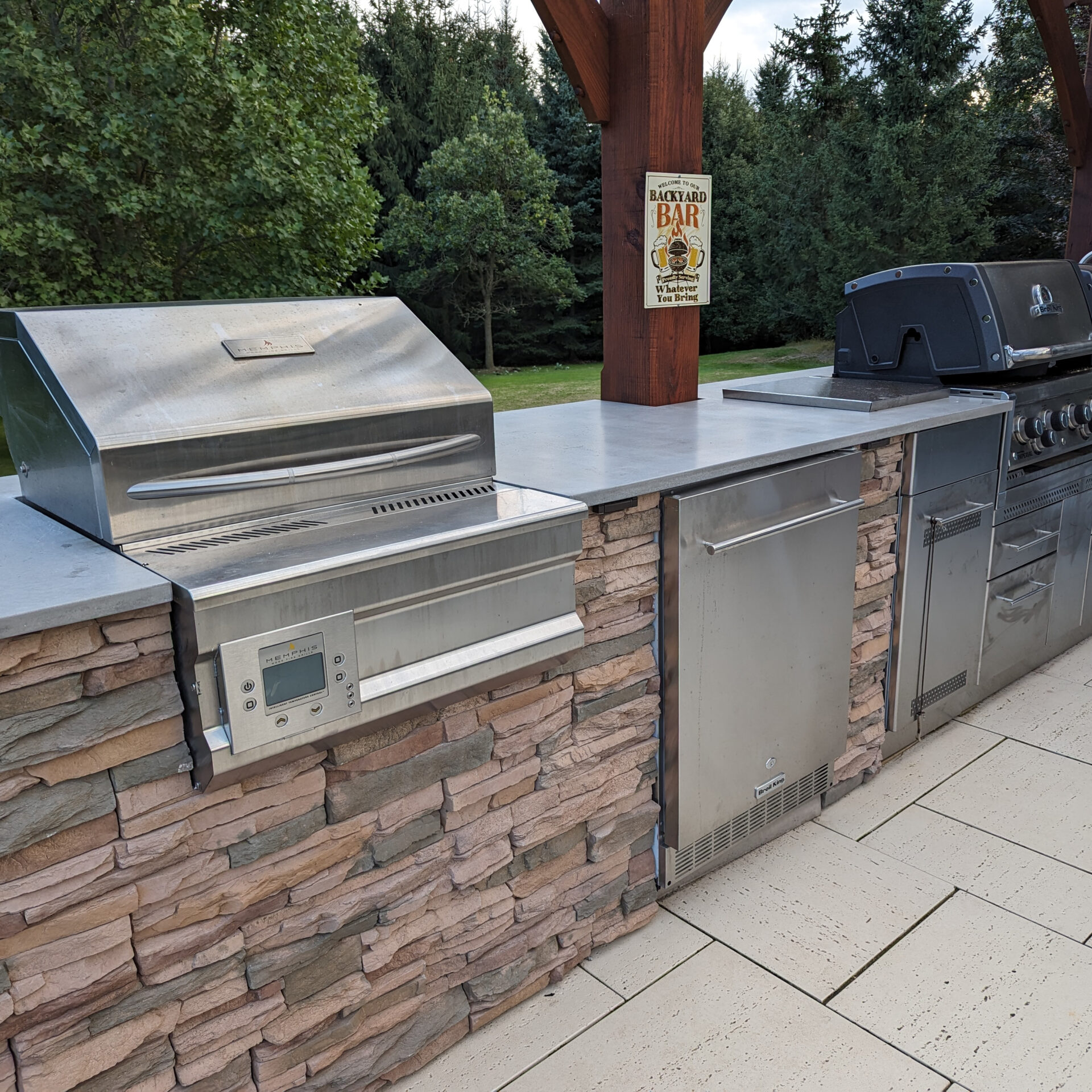 Outdoor kitchen with stainless steel appliances and stone finish. "Backyard Bar" sign on wooden post. Lush green trees in the background.