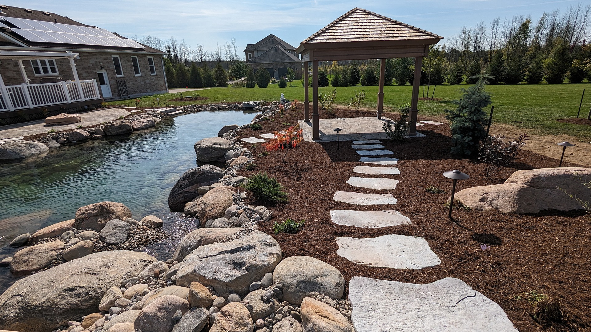 A serene backyard with a pond, stone path, and gazebo. Solar panels are installed on the house. Green lawn and trees surround.