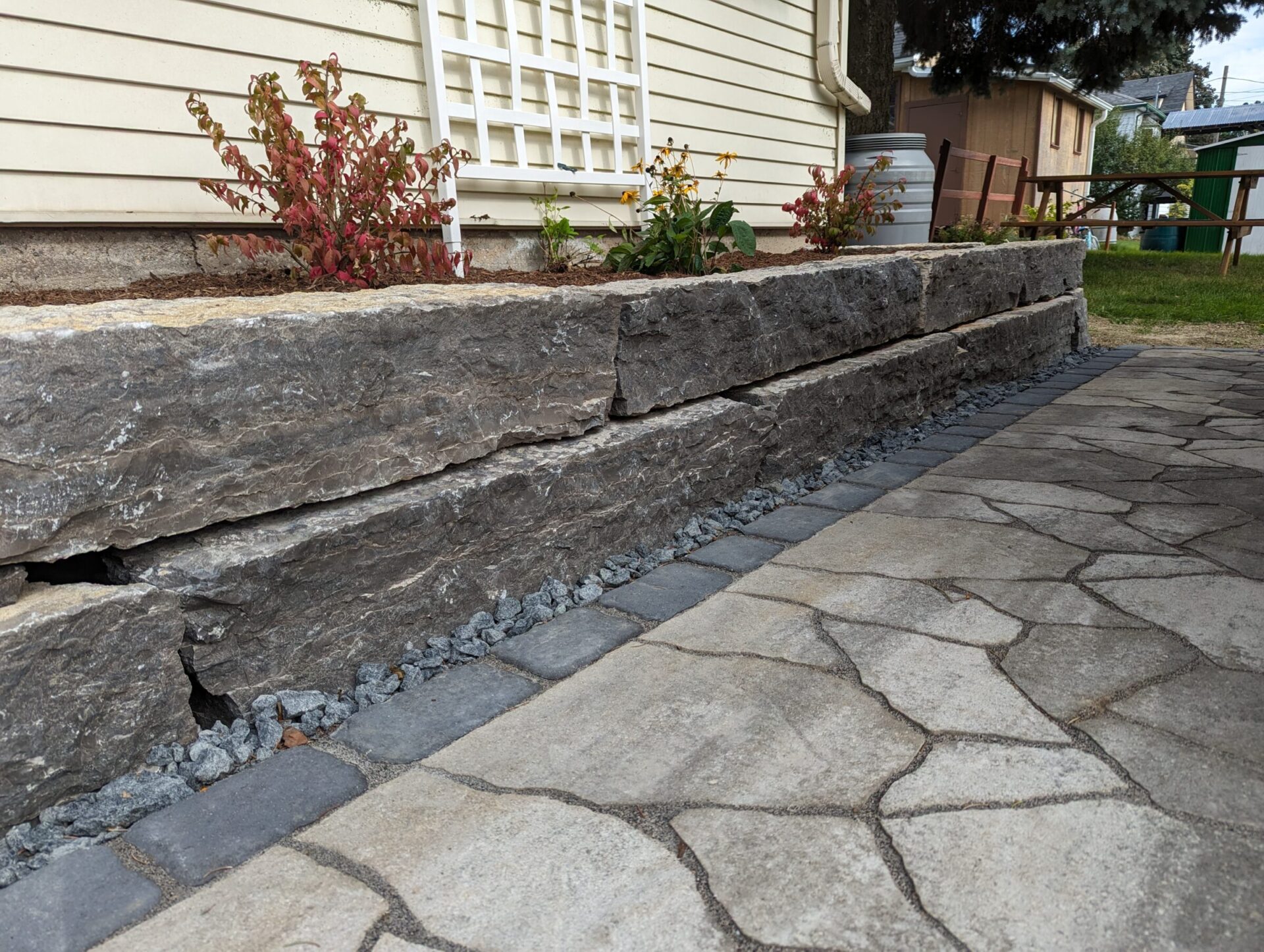 Stone retaining wall with small plants against a house. Pathway made of irregular stone slabs. Backyard setting with grass and fence.