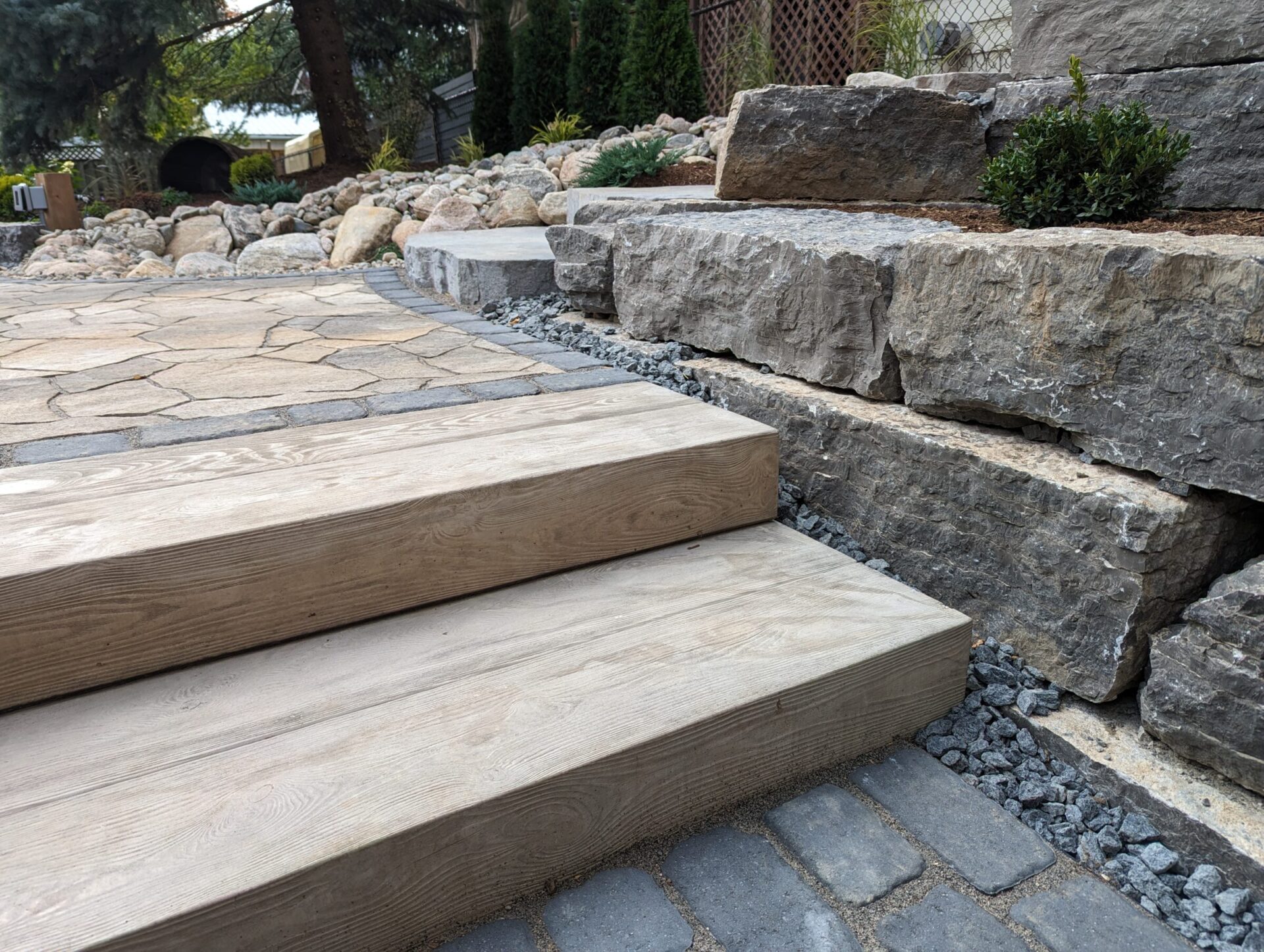 Wooden steps lead to a stone patio, surrounded by rocks and greenery in a landscaped garden. The scene is calm and natural.