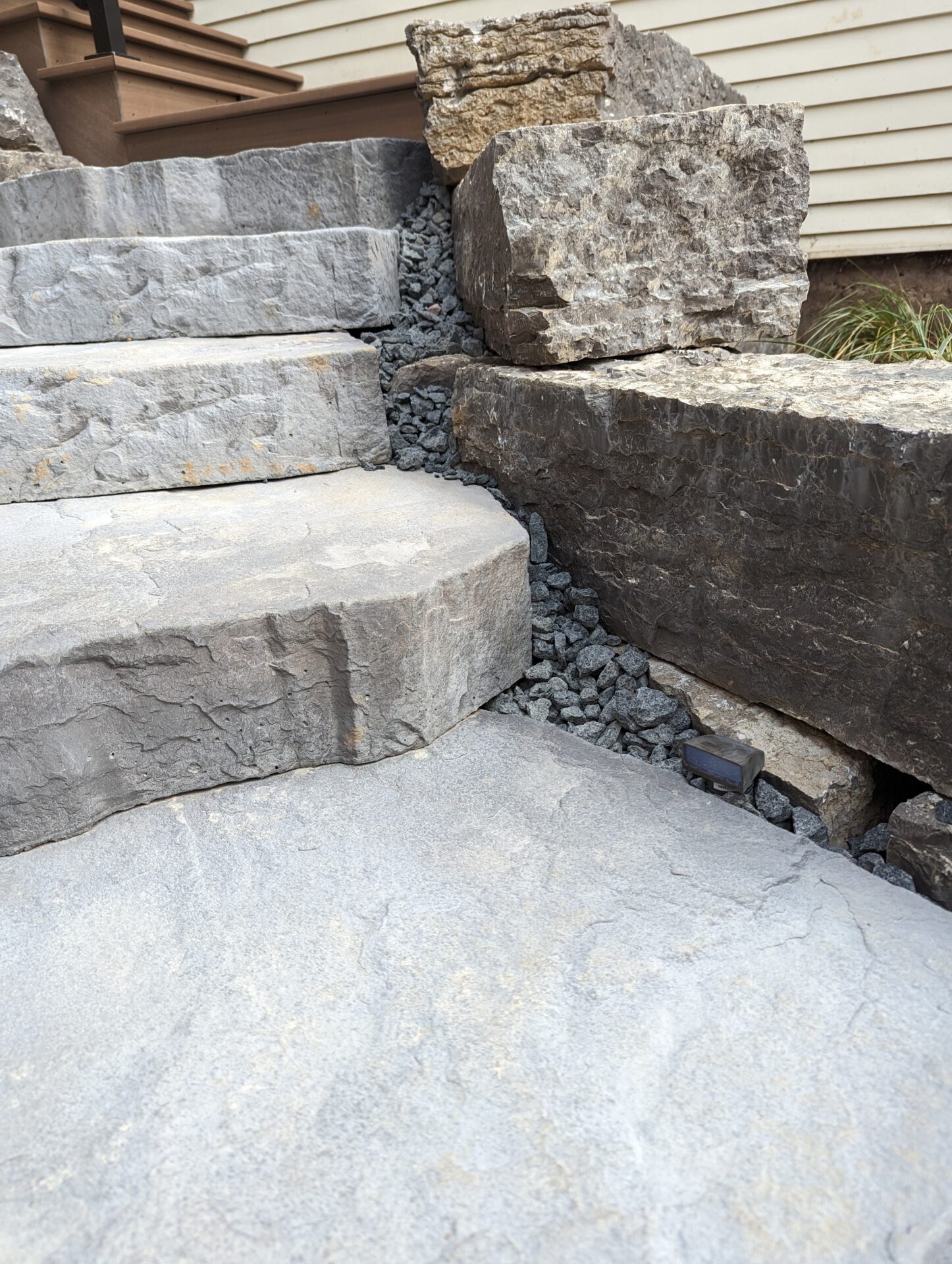 Stone stairs and large rocks create a rustic outdoor pathway. Crushed gravel fills gaps between stones, leading up toward a wooden structure.