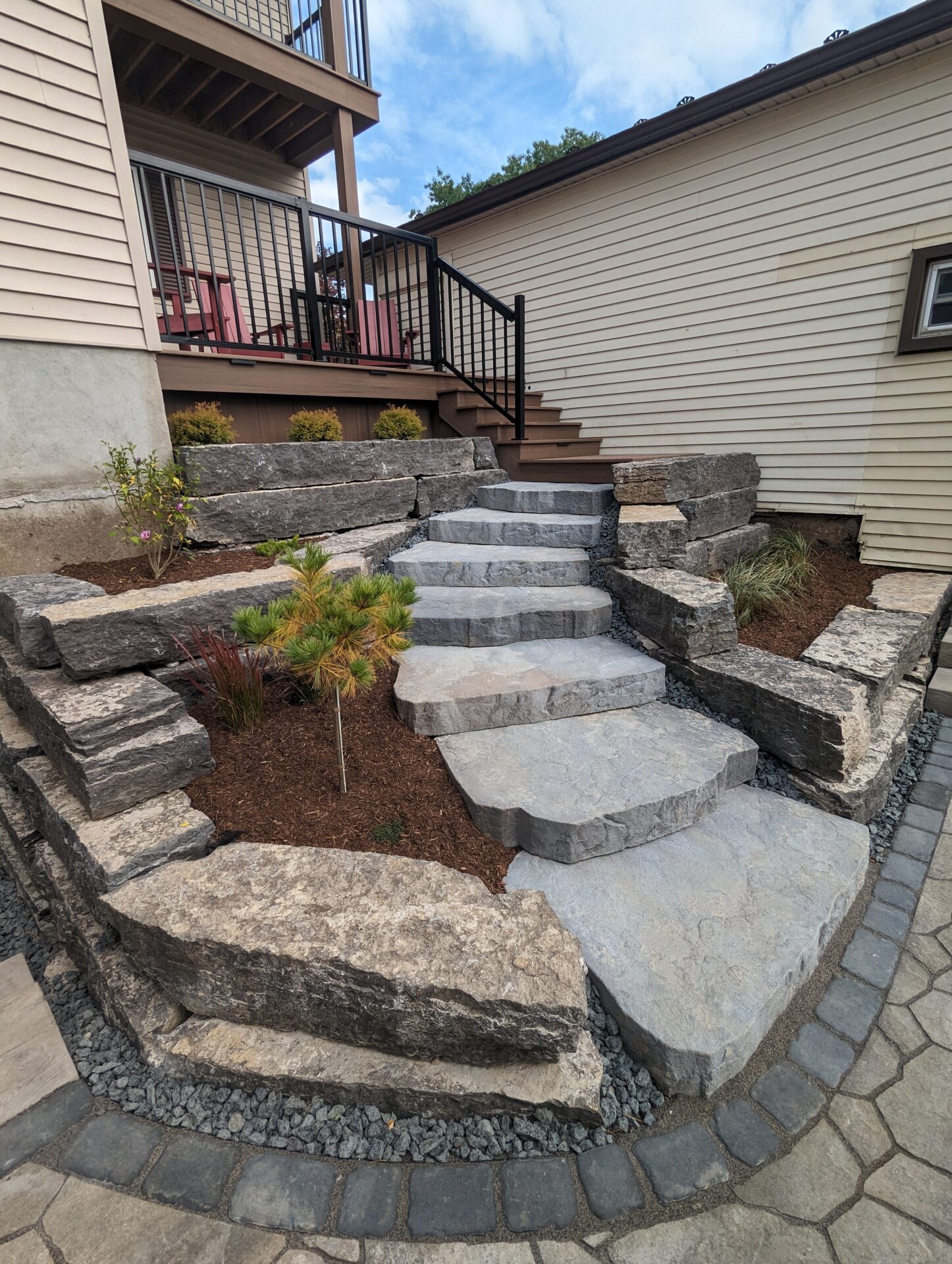 Stone steps lead to a wooden deck with chairs, bordered by landscaped rocks and plants, against light-colored house siding under a blue sky.