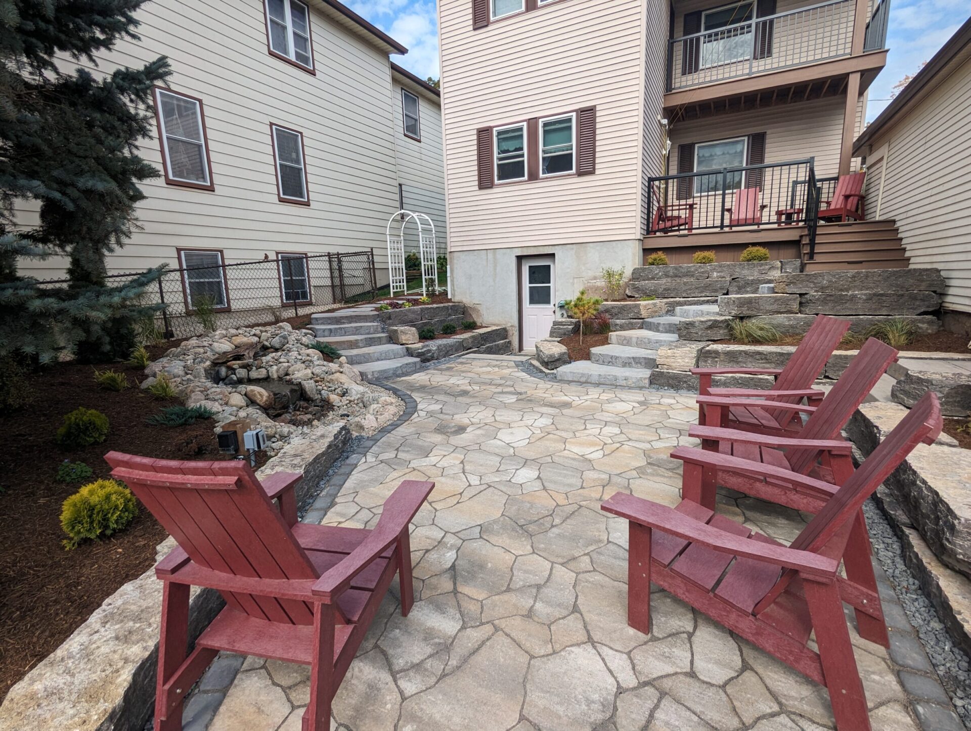 Patio area with red chairs, stone pathways, landscaped garden, small waterfall feature, and two adjacent multi-story houses with windows and balconies.