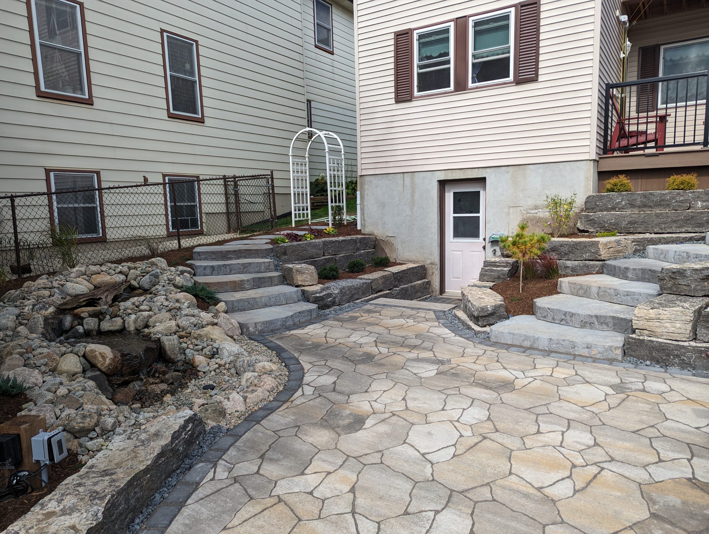 A landscaped backyard with stone steps, a small rock waterfall, and a white arbor near a two-story building's entrance.