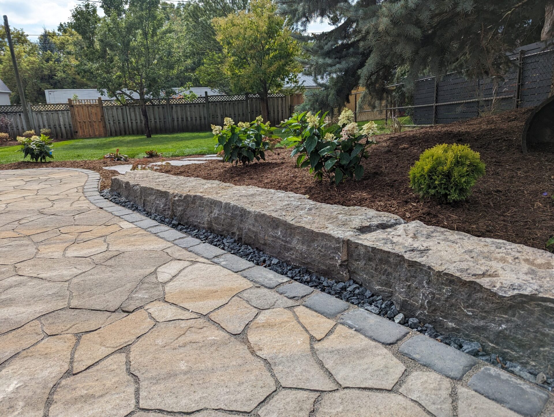 A landscaped backyard features a stone patio, bordered by a rock wall with mulch and plants, adjacent to a fenced lawn area.