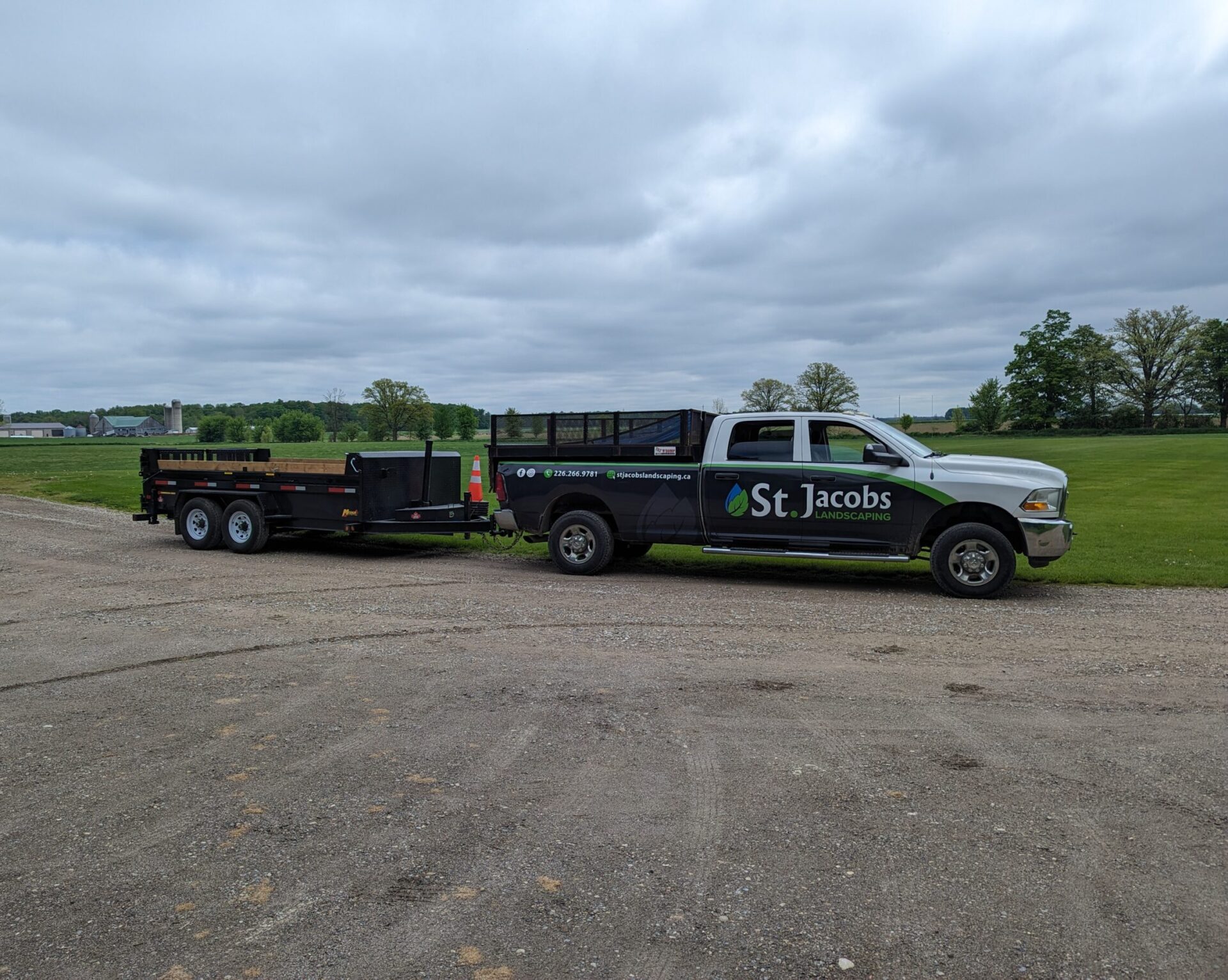 A truck with "St. Jacobs Landscaping" signage tows a trailer on a gravel path, surrounded by green fields and distant farm structures.