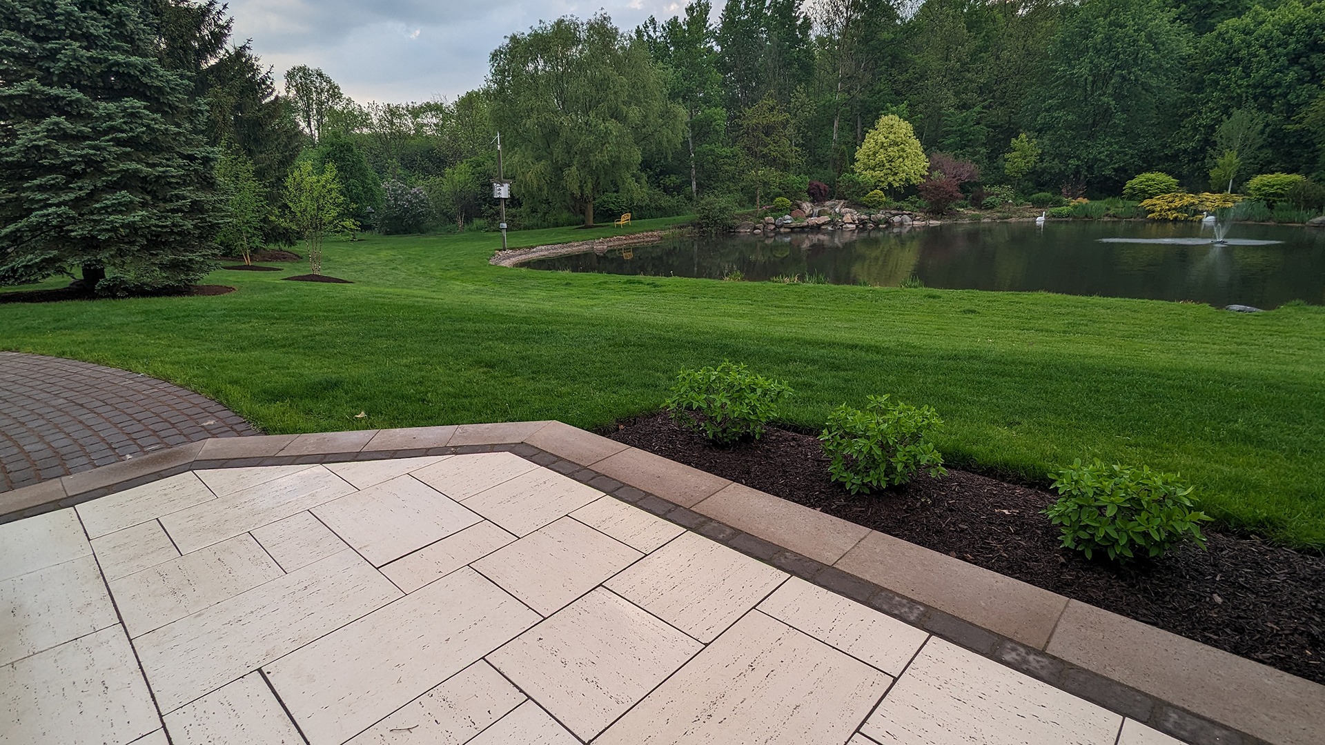 A serene park scene with a pond, fountain, lush trees, and a paved walkway. Peaceful and inviting atmosphere for relaxation.